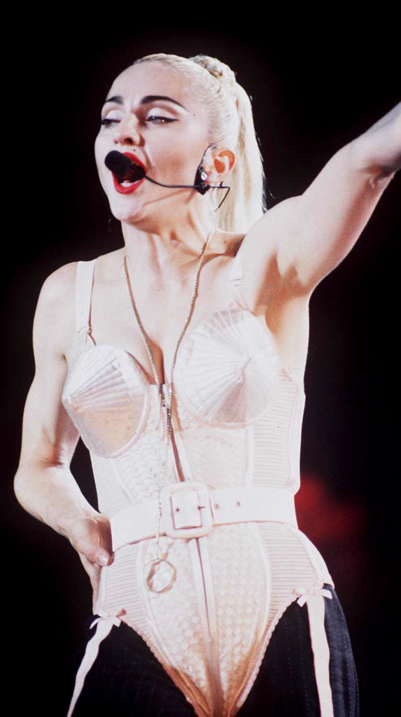 Madonna on stage, wearing a cone bra corset and a headset microphone, performing energetically with one arm raised