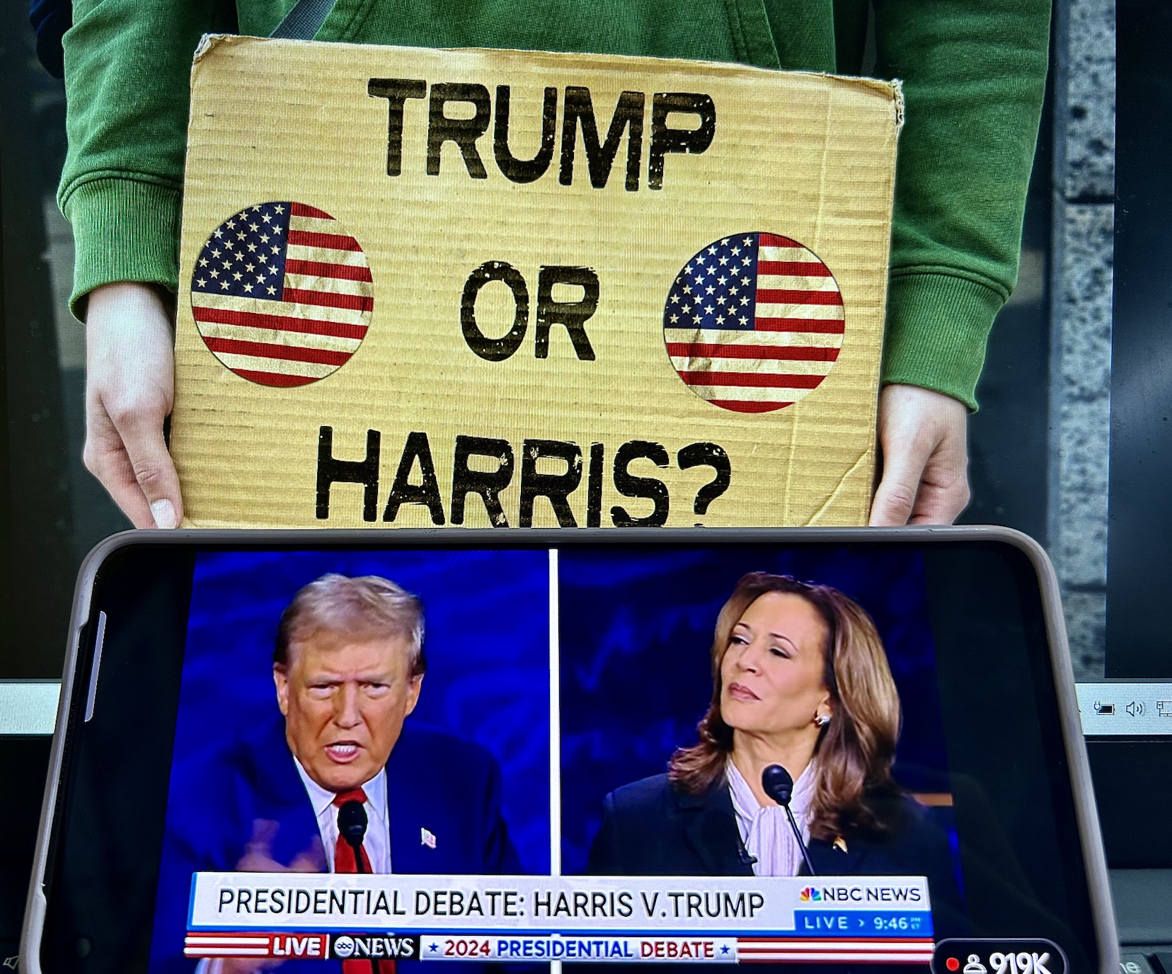 Person holding a sign reading "TRUMP OR HARRIS?" with images of American flags, while a phone screen shows Donald Trump and Kamala Harris during a debate