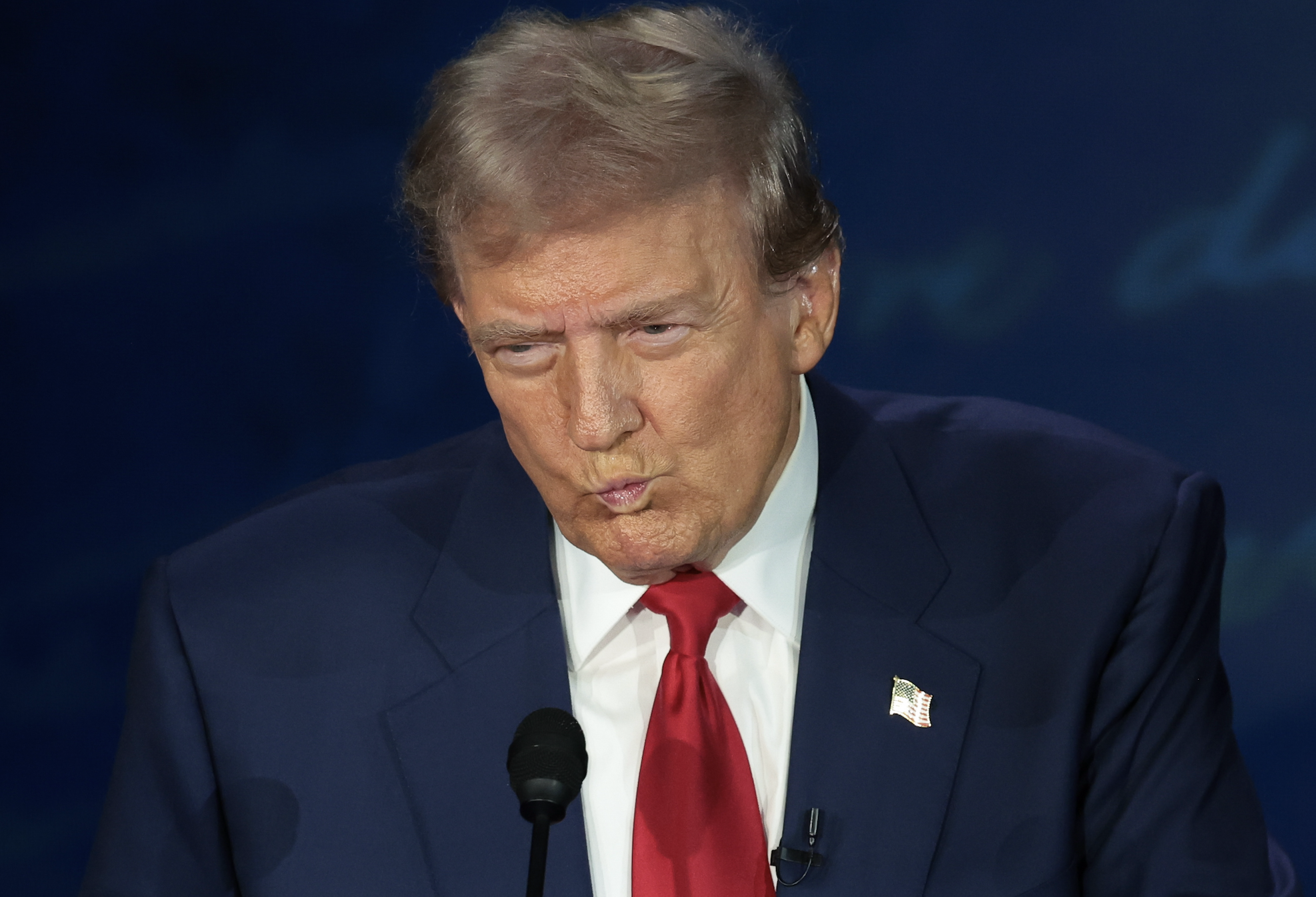 Donald Trump at a speaking event, wearing a suit and tie, standing at a podium with a serious expression