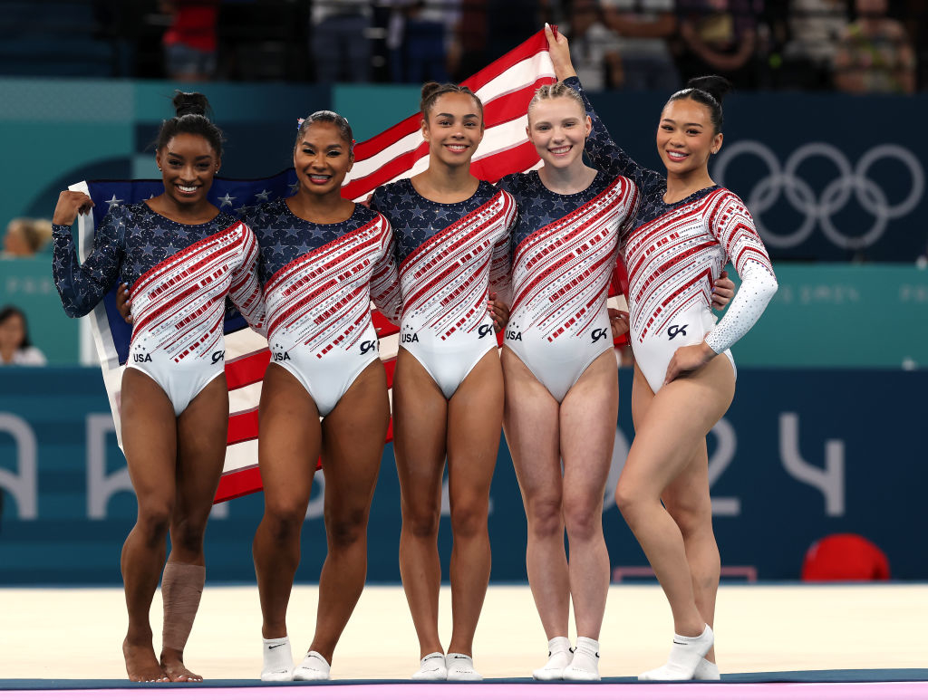Simone Biles, Jordan Chiles, Hezly Rivera, Jade Carey, and Sunisa Lee in Team USA gymnastics uniforms holding an American flag at the Olympics