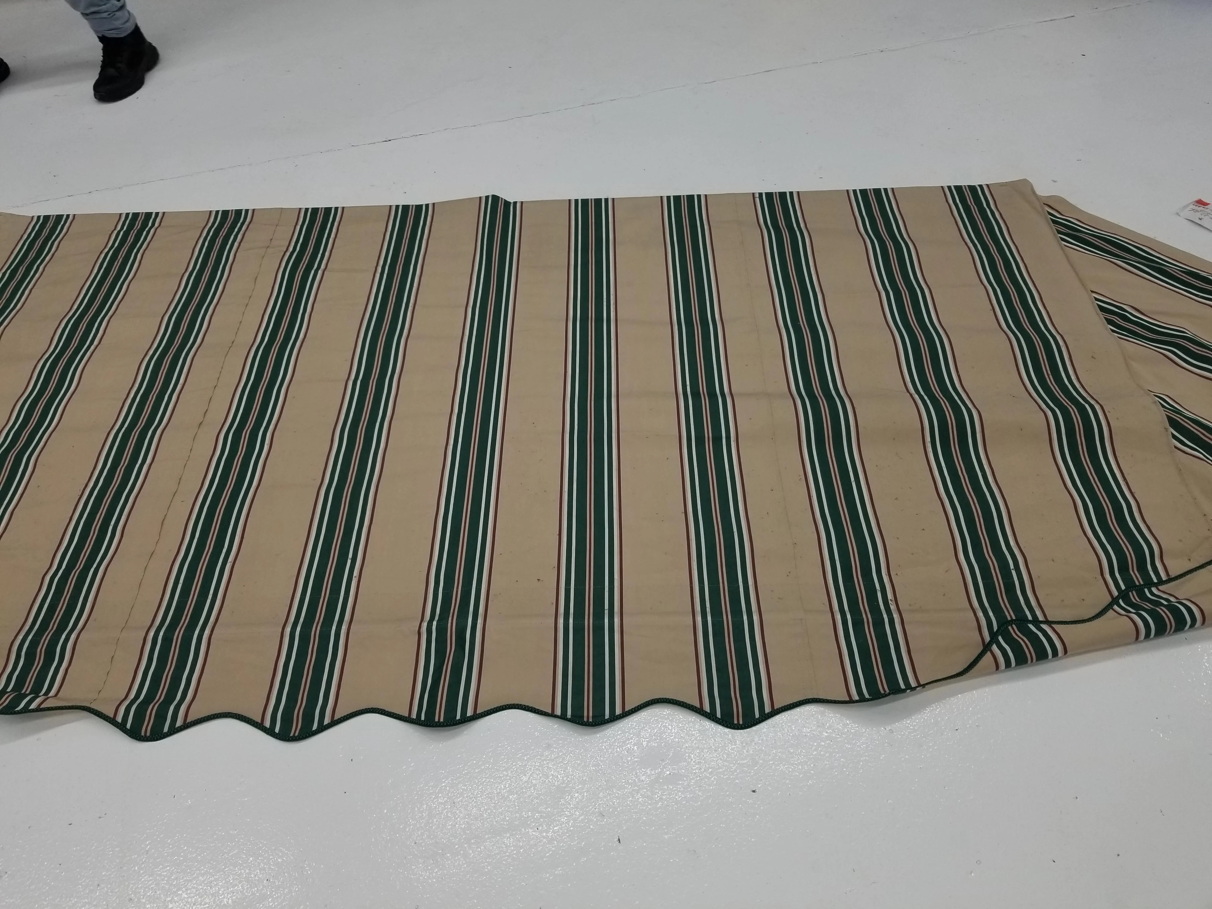 A large, striped outdoor mat is spread flat on a white floor. Only part of a person's legs is visible at the top left of the image