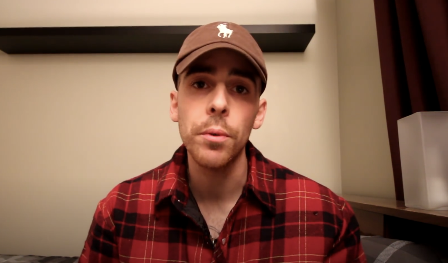 A man wearing a brown baseball cap and a red plaid flannel shirt is sitting in a room with a black shelf on the wall behind him