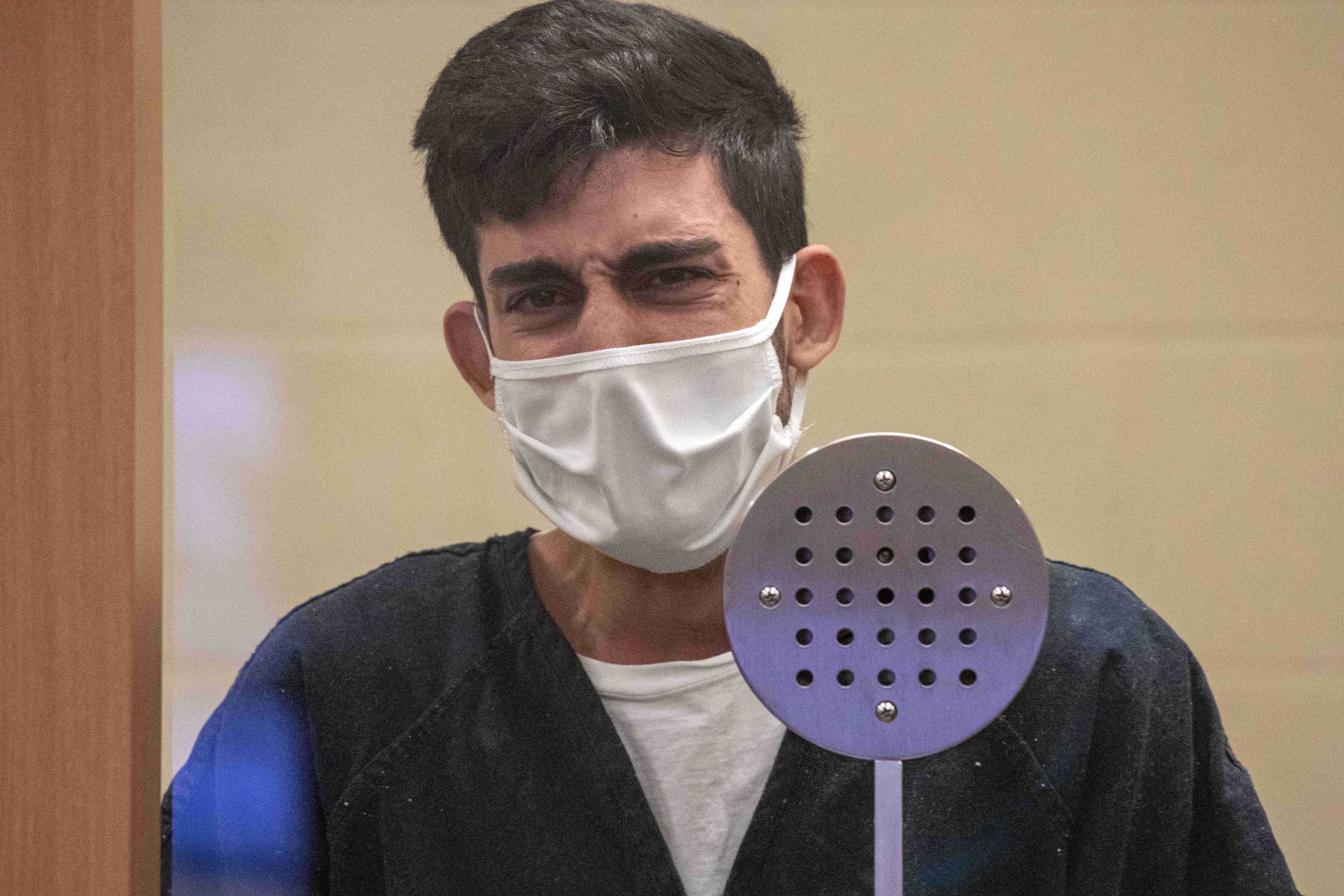 A man in a surgical mask is standing behind a glass barrier in what appears to be a courtroom, speaking into a microphone