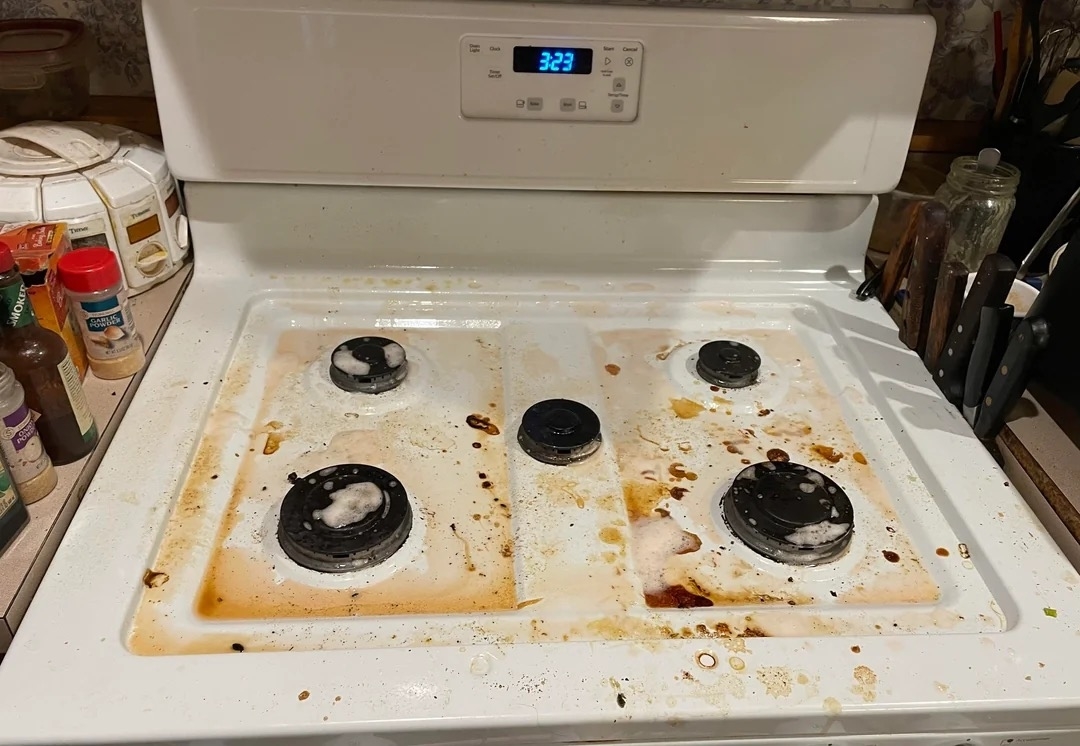 A white kitchen stove with four burners is visibly dirty with spills and stains. Cooking utensils and spices are seen on the counter. The clock displays 3:23