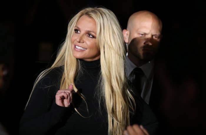 Britney Spears smiles and holds sunglasses, wearing a high-neck dark top. A security guard stands in the background