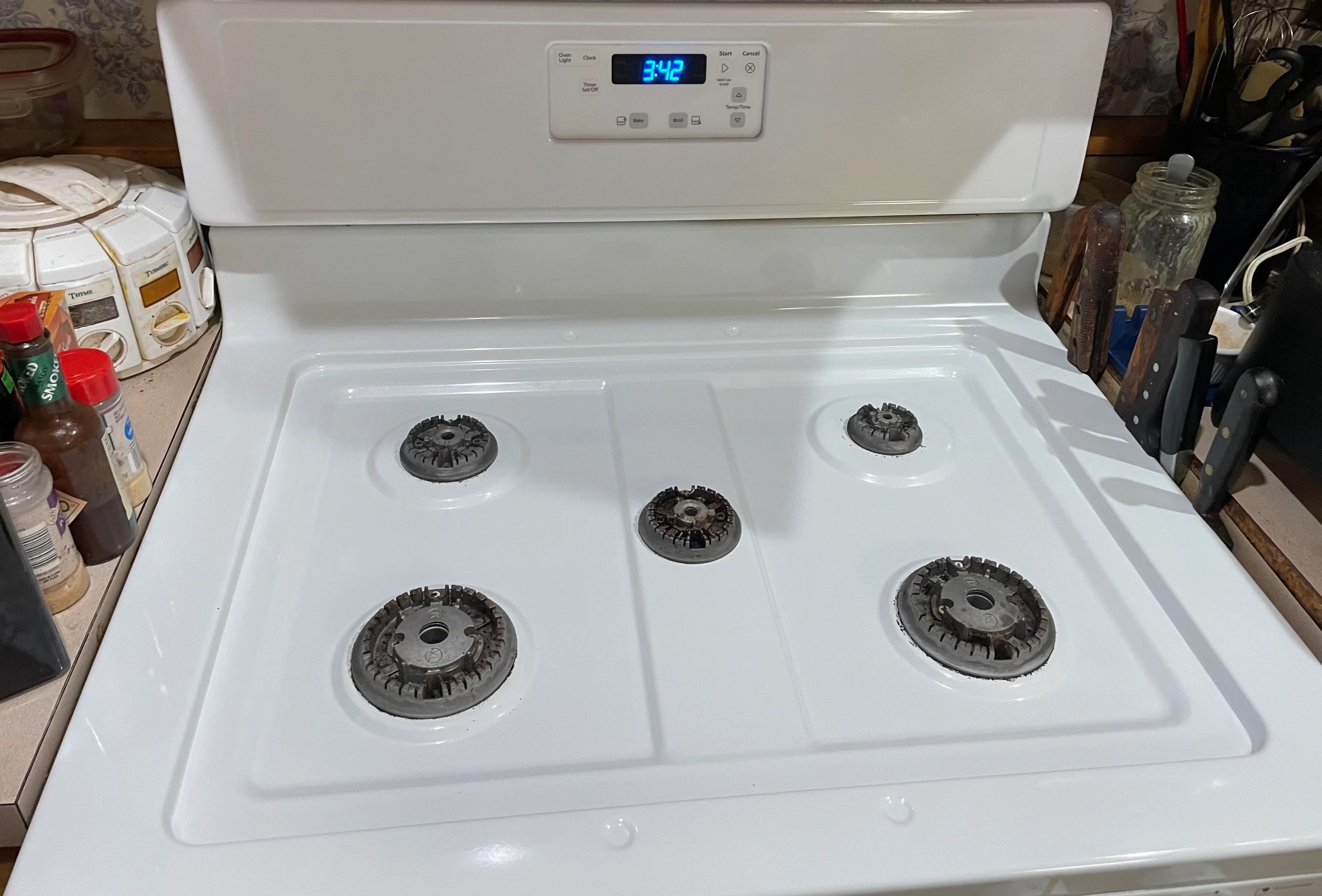 Gas stovetop with five burners, one large center burner and four smaller surrounding burners. The clock display shows 3:42. Kitchen utensils and spices are visible in the background