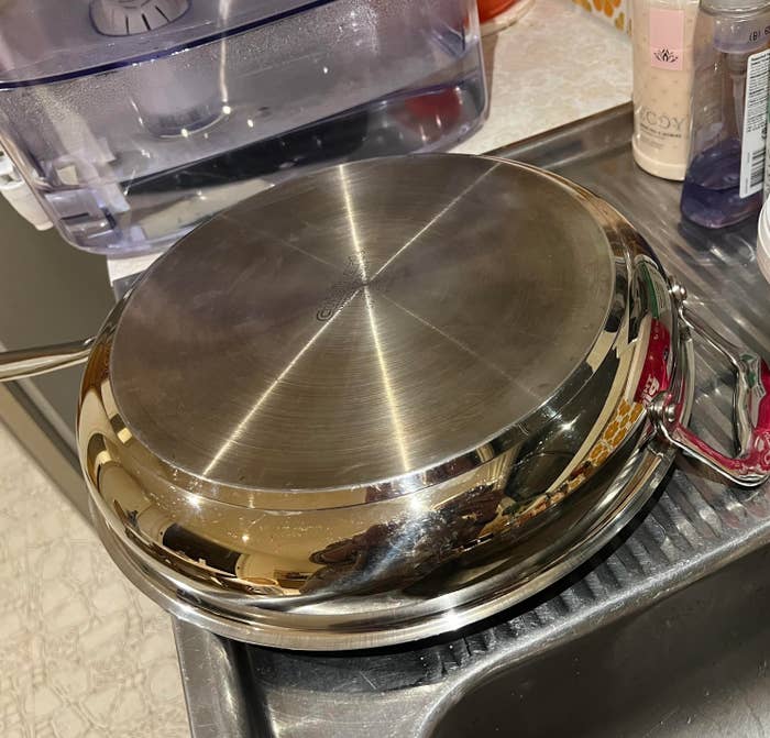 A stainless steel pan is upside down, drying on a kitchen sink rack. Cleaning products and a water filter are visible in the background