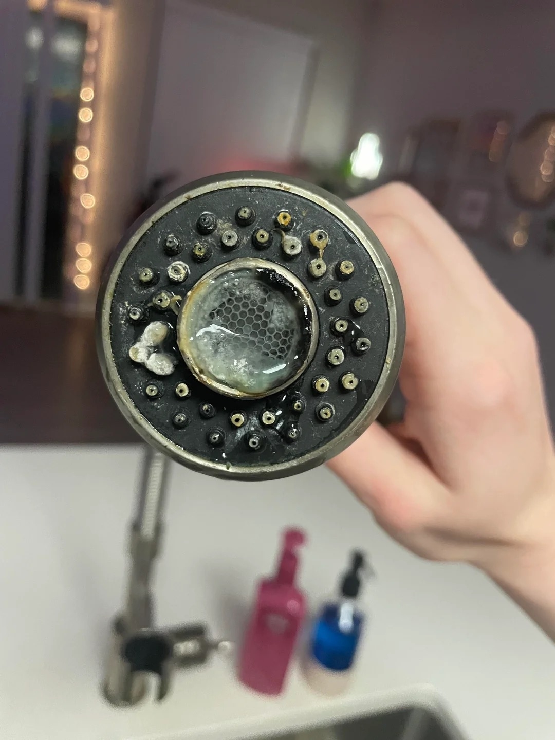 Close-up of a hand holding a showerhead with mineral buildup and grime. Kitchen counter and fixtures are visible in the background