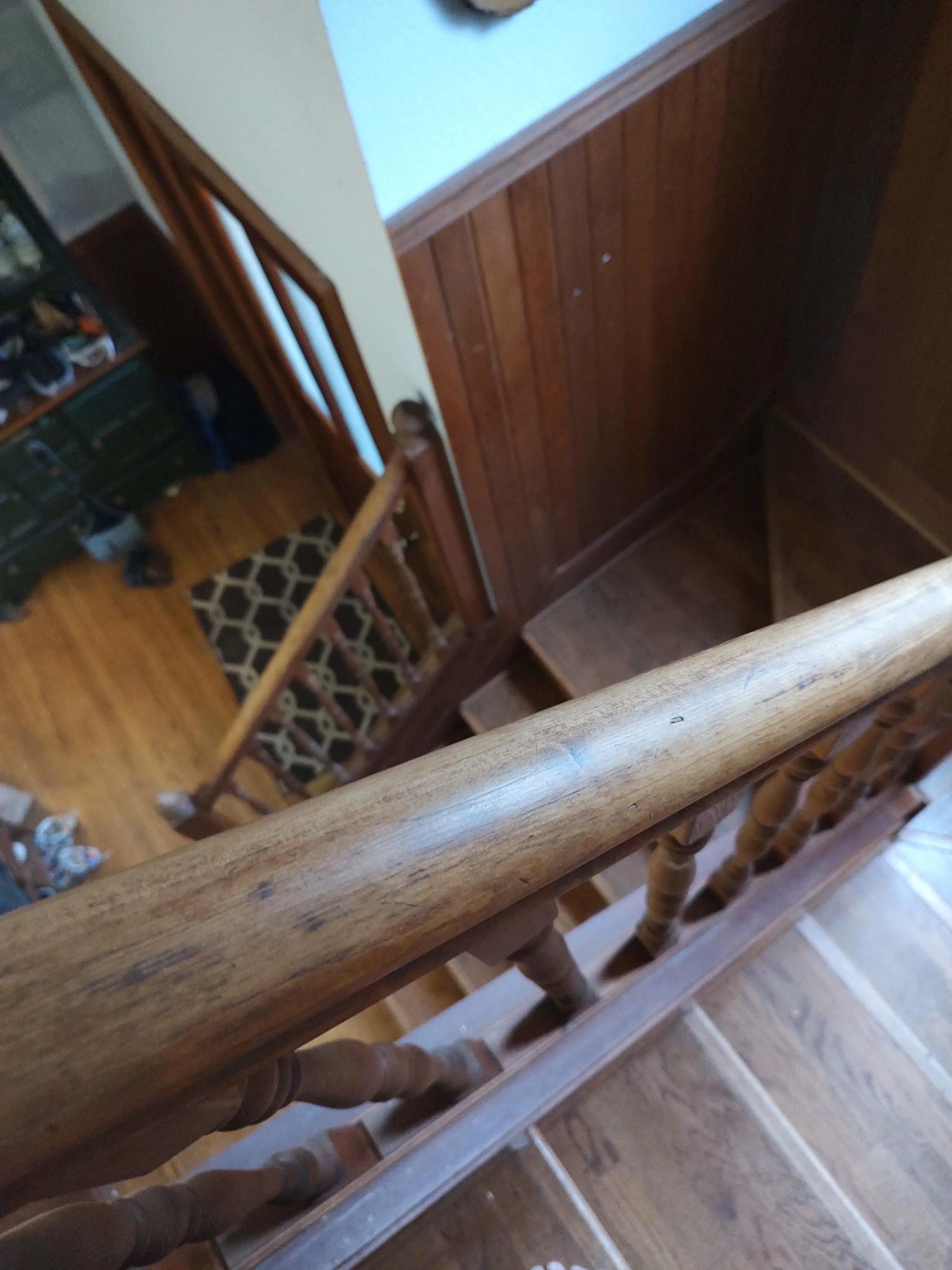 View from the top of a wooden staircase looking down, with wooden handrails and steps. A patterned rug and a cabinet with shoes are visible at the bottom