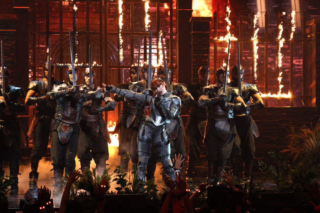 Chappell Roan, dressed in knight armor, performs on stage with background dancers in armor, with fire effects in the background