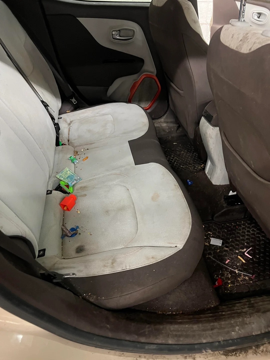 A dirty car interior with scattered trash and toys on the back seat and floor. The surfaces are stained and the car appears untidy. No persons are present