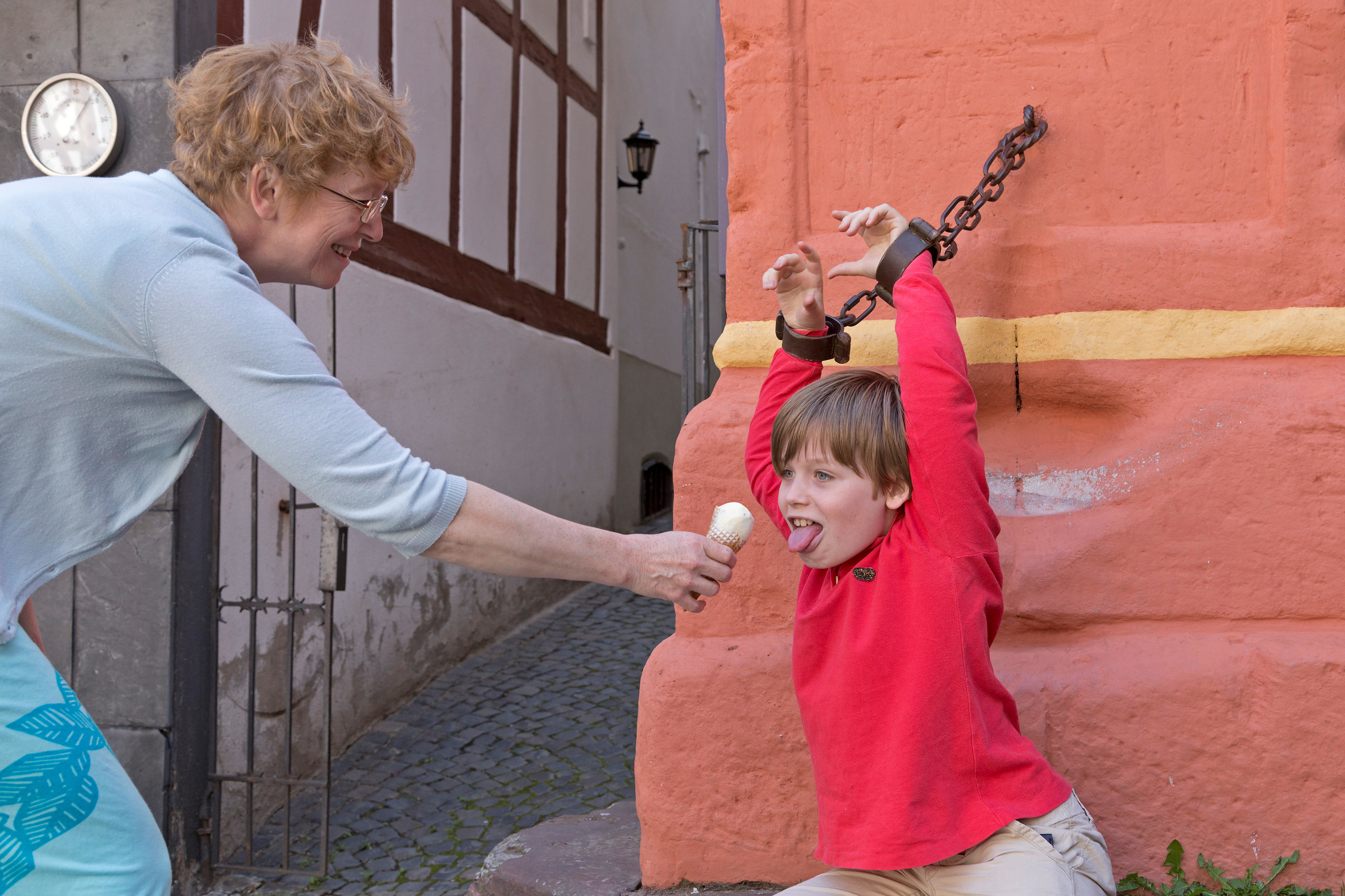 A woman playfully offers an ice cream cone to a smiling young boy who is pretending to be chained to a wall for fun