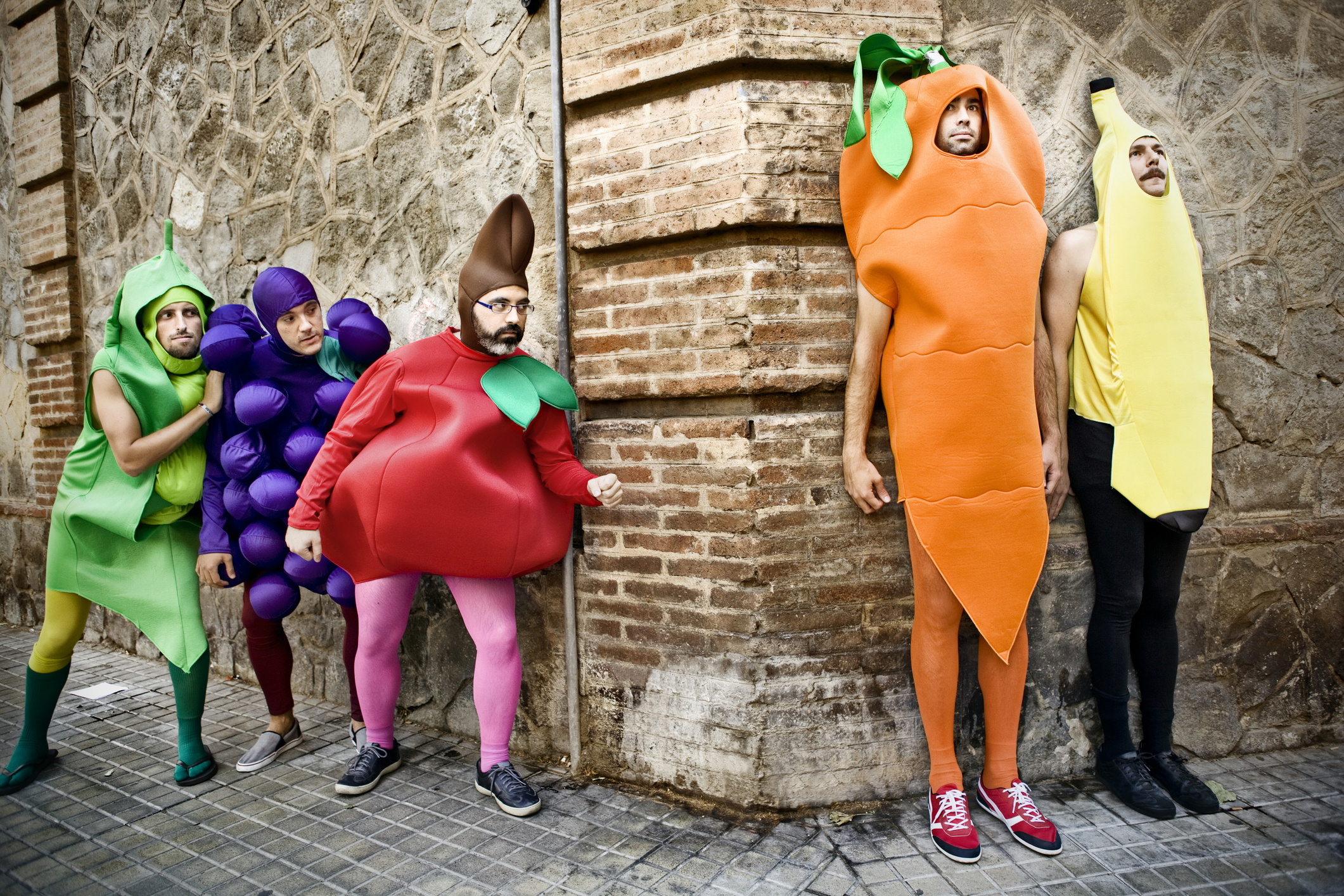 Four people dressed as fruits and vegetables (pea pod, grape bunch, chili pepper, carrot, and banana) pose and smile near a brick wall. Names unknown