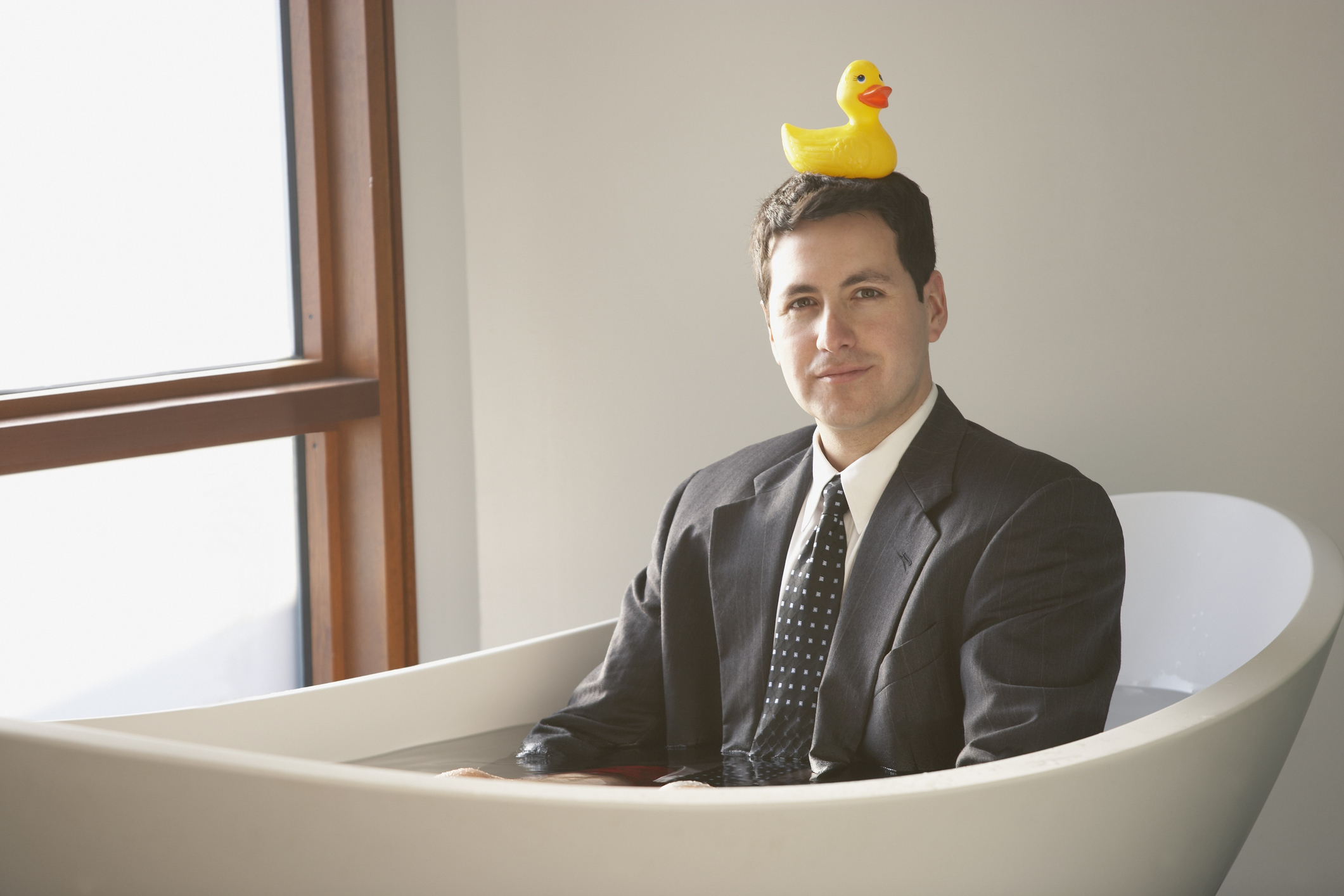 Man in a suit sitting in a bathtub with a rubber duck on his head, looking at the camera