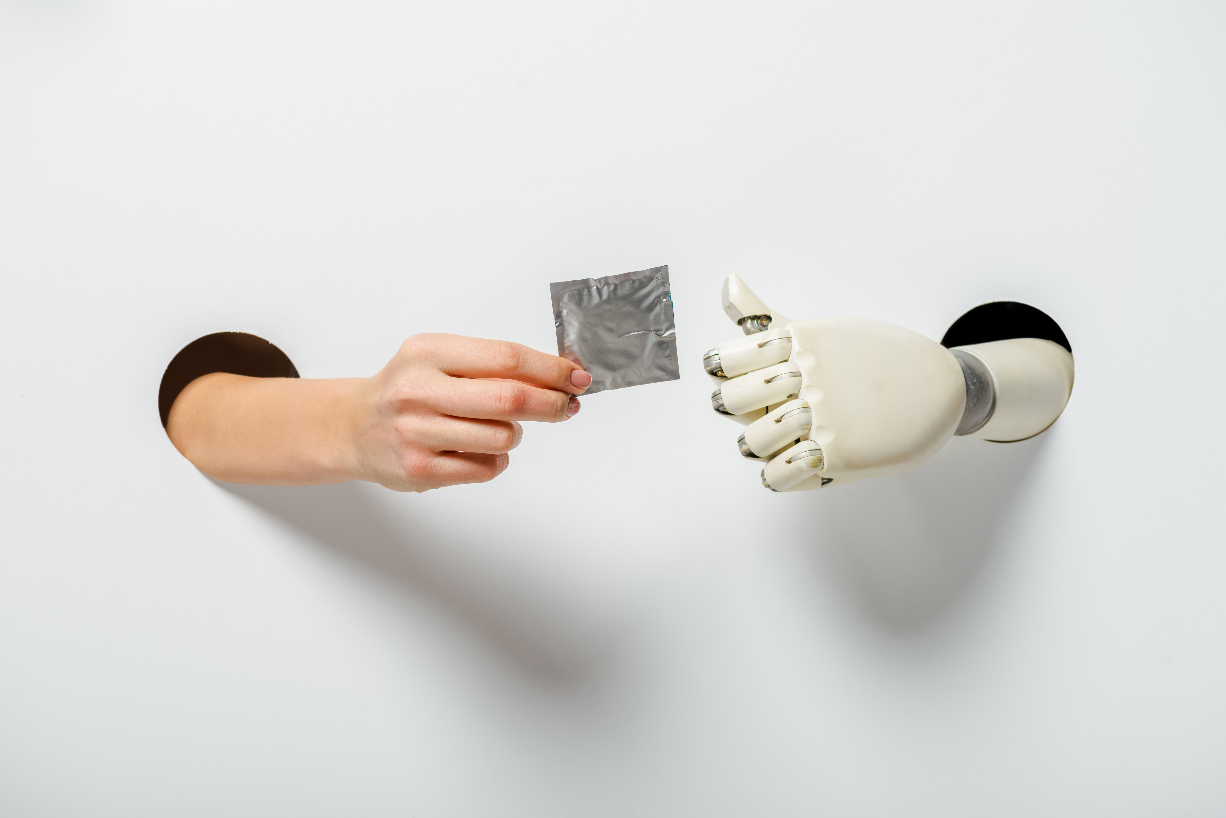 A human hand and a robotic hand, both extending through holes in a white background, with the human hand giving a condom to the robotic hand