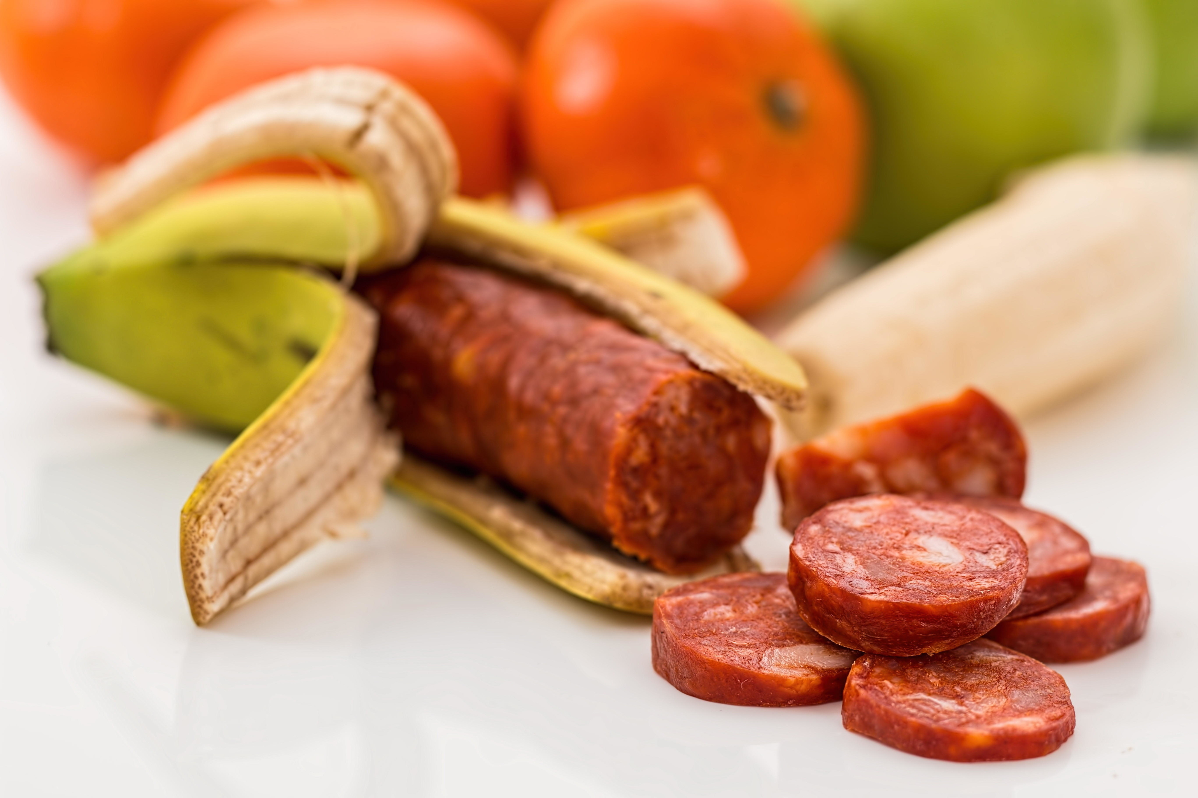 A banana peel holds a sausage, with several sausage slices in front. Whole fruits, such as mandarins and another banana, are in the background