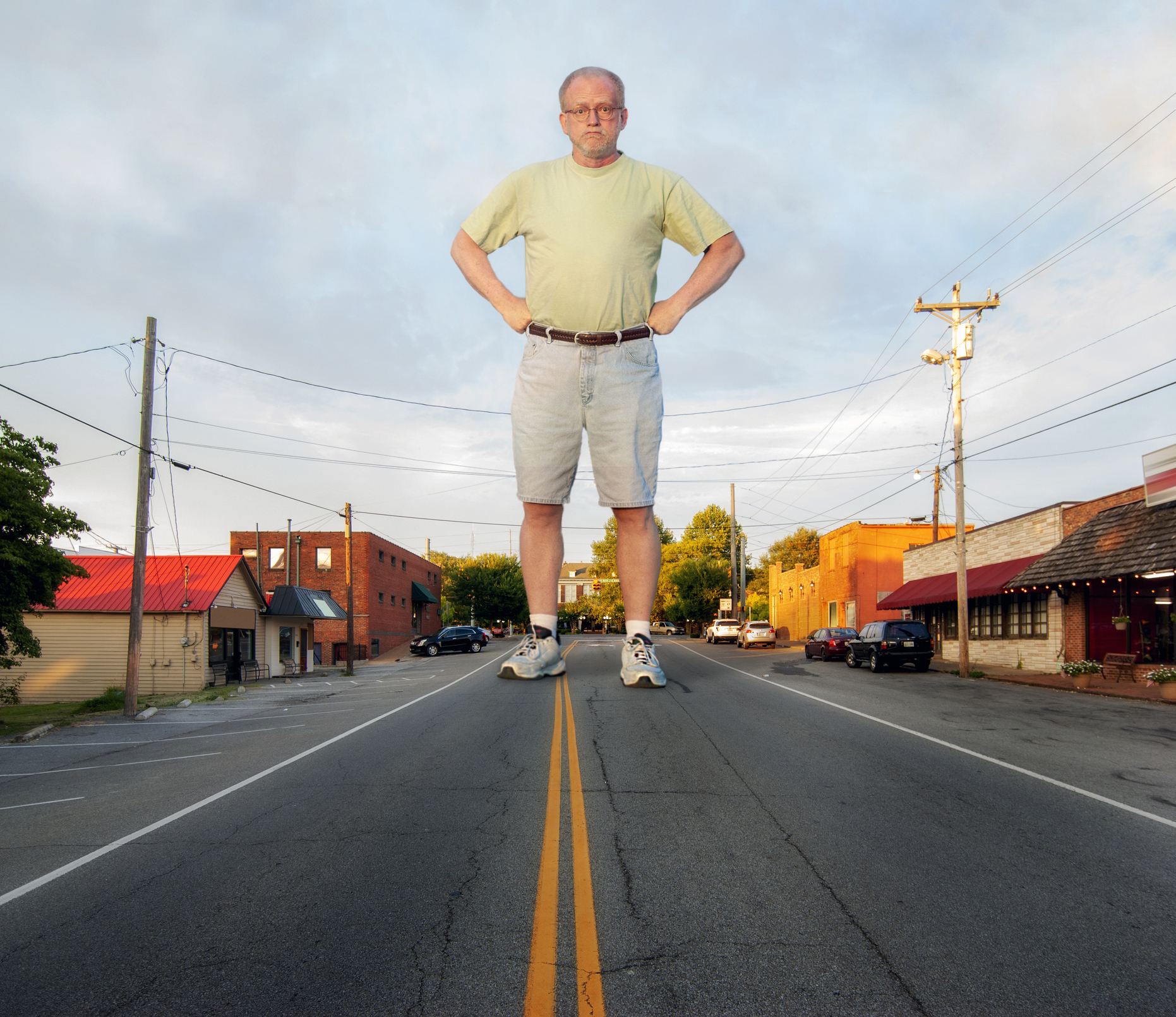 A man stands in the middle of a road, appearing disproportionately large compared to the surrounding buildings and cars. Hands are on his hips