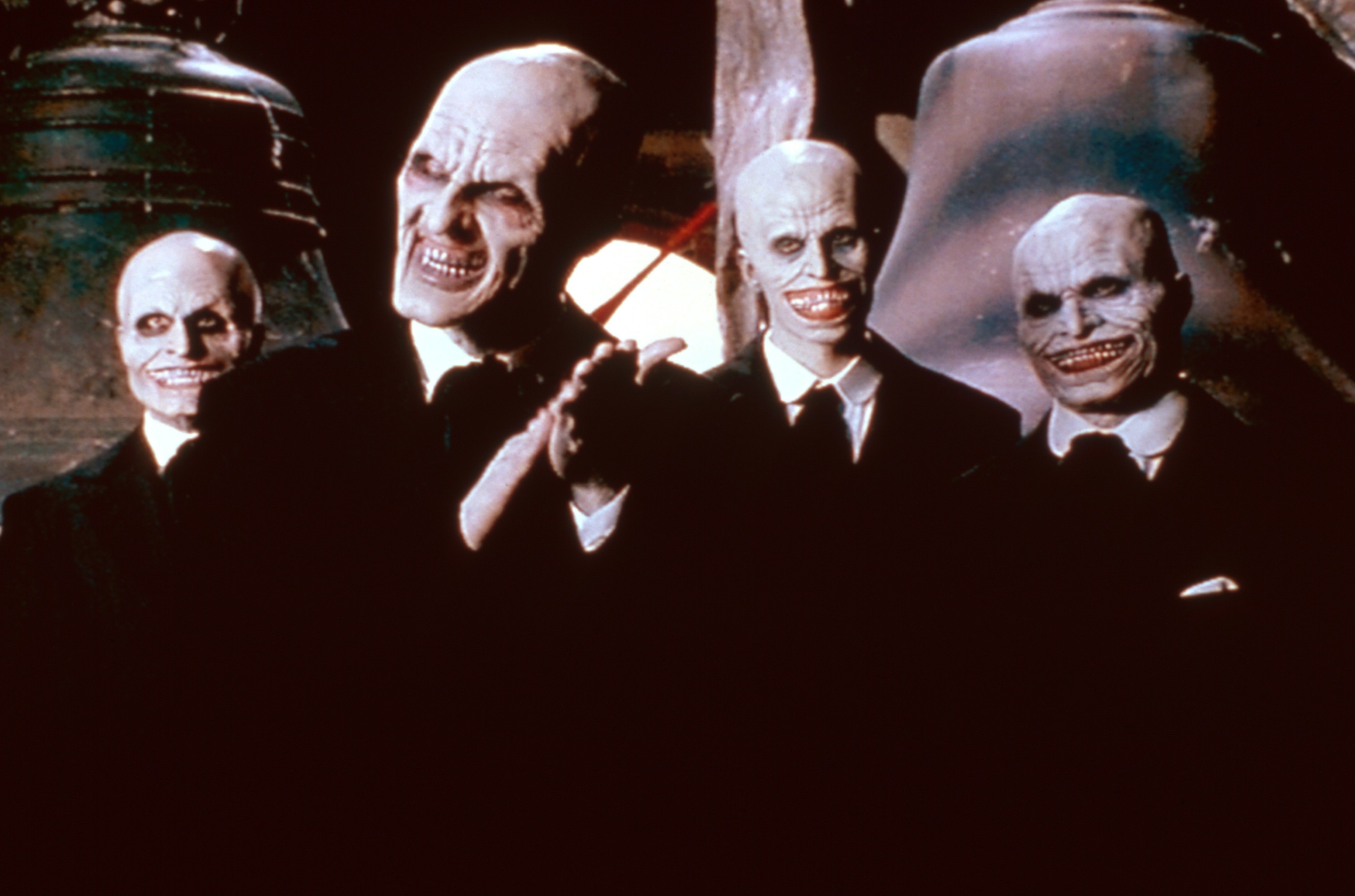 Four creatures with bald heads, pale skin, sharp teeth, and wide grins wearing business suits, standing in a dimly lit setting from Buffy the vampire slayer