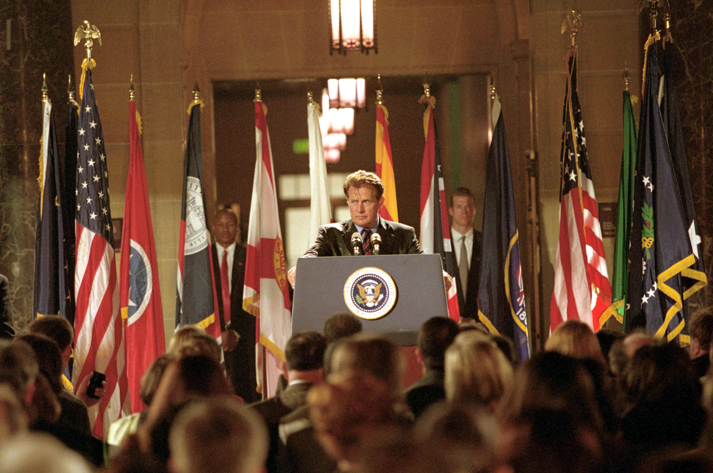 Martin Sheen , standing at a podium with the presidential seal, addresses a crowd in a formal setting surrounded by multiple state and national flags