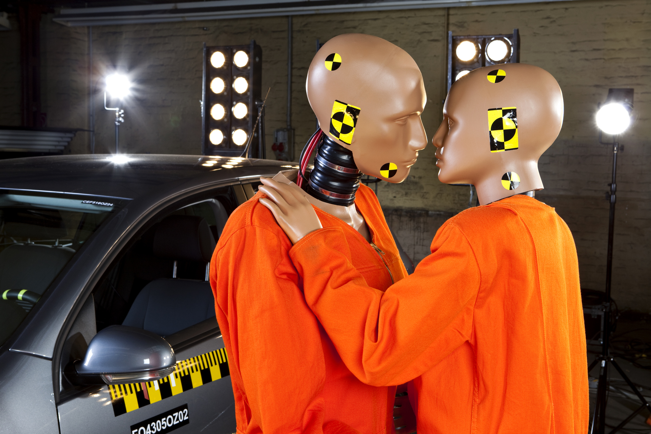 Two crash test dummies wearing orange suits stand close together, facing each other next to a test car with bright lights in the background