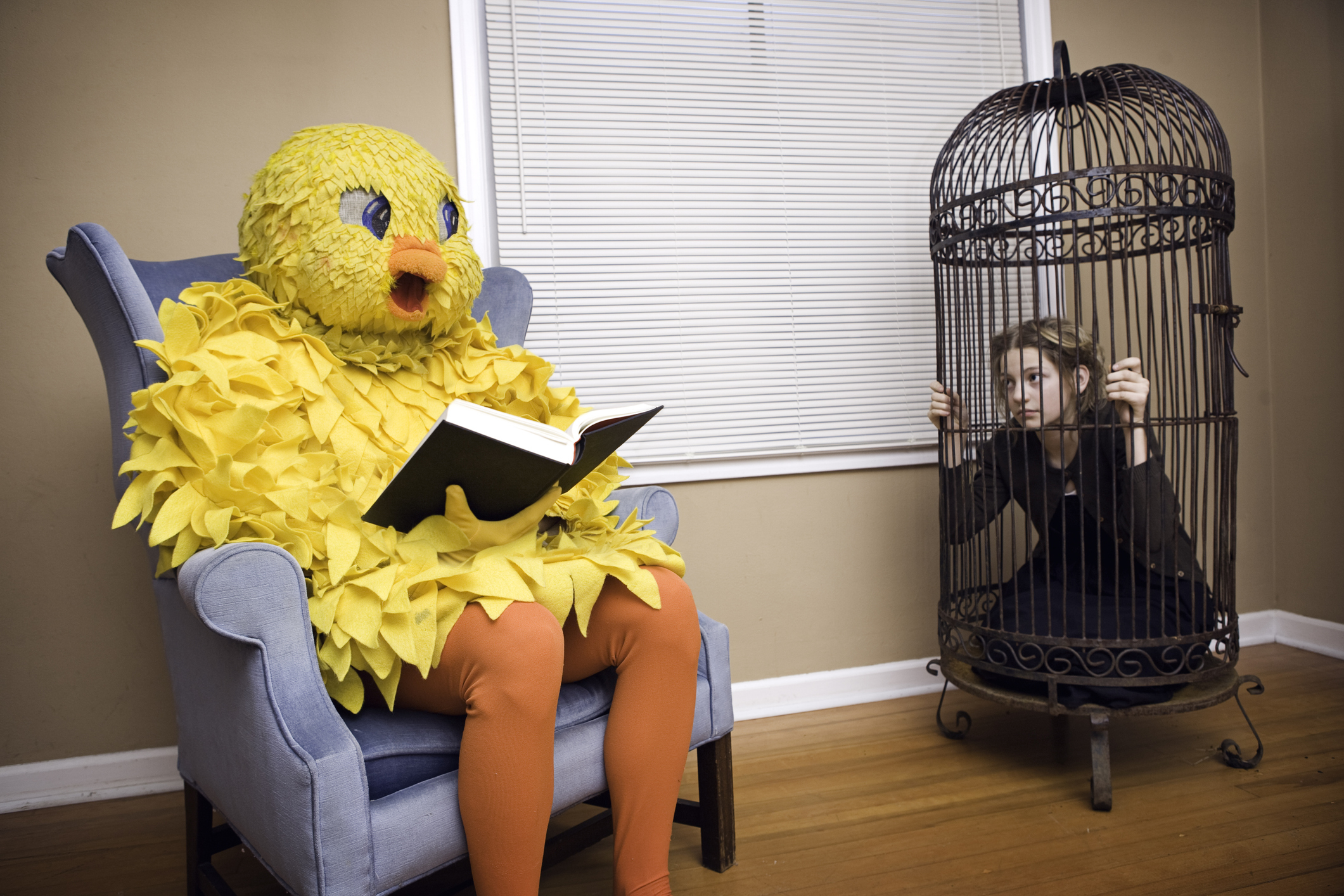 Person in a bird costume reading a book on an armchair while another person is sitting inside a birdcage looking at them. Names unknown