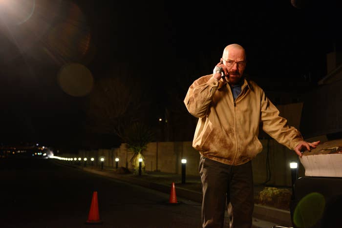 Bryan Cranston as Walter White in Breaking Bad stands by a car at night, talking on his phone, wearing a beige jacket. Cones and lights visible in the background