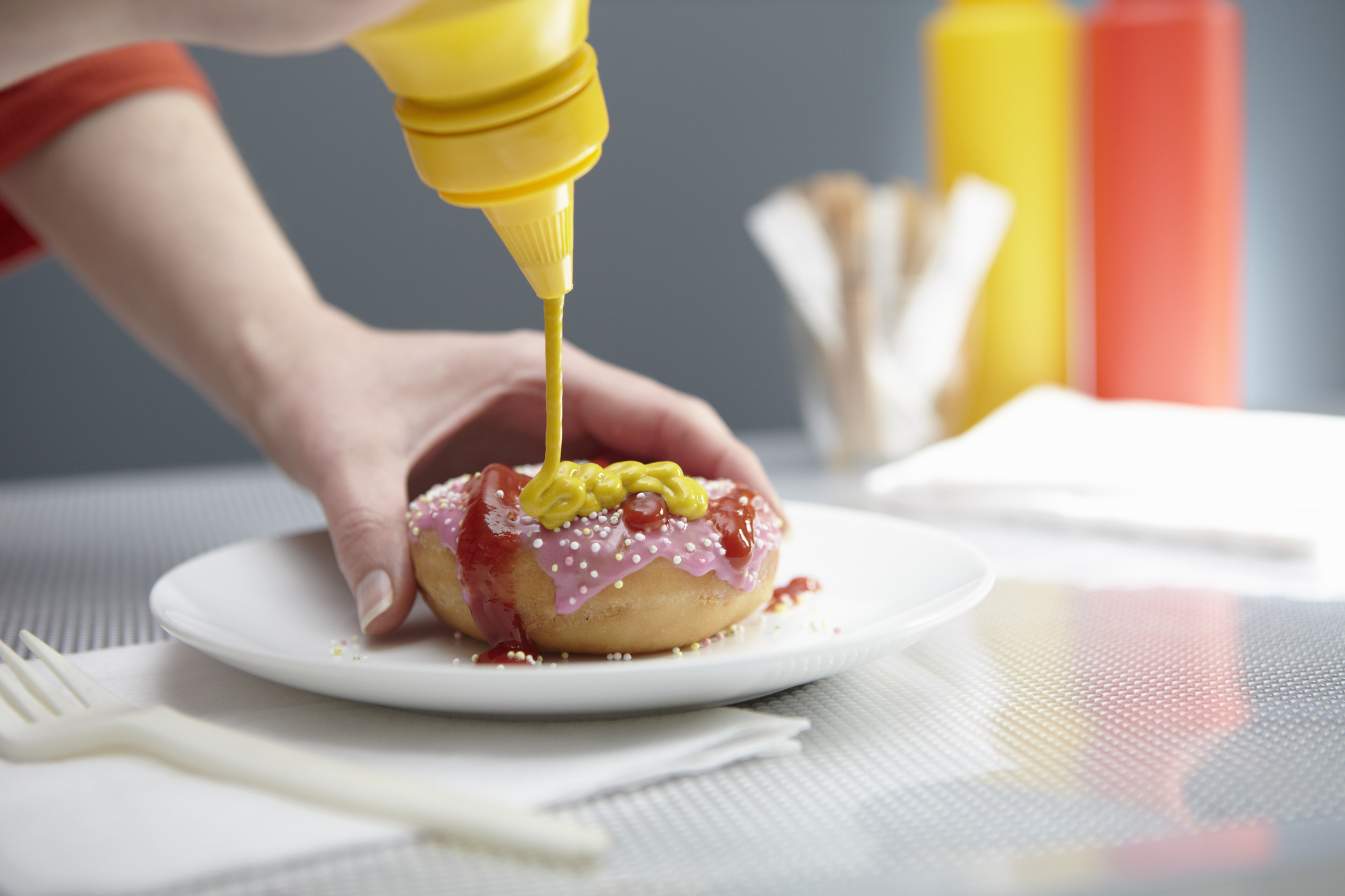 A person is pouring yellow mustard onto a colorful donut on a white plate, with blurred condiments and napkins in the background
