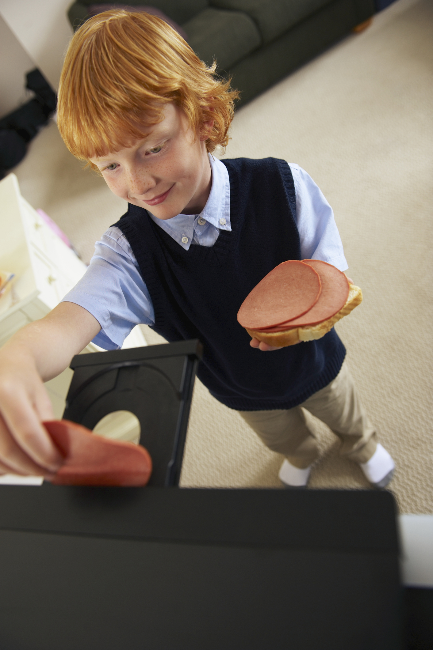 A young red-haired boy mistakenly inserts a slice of bologna into a DVD player while holding a sandwich with bologna in the other hand