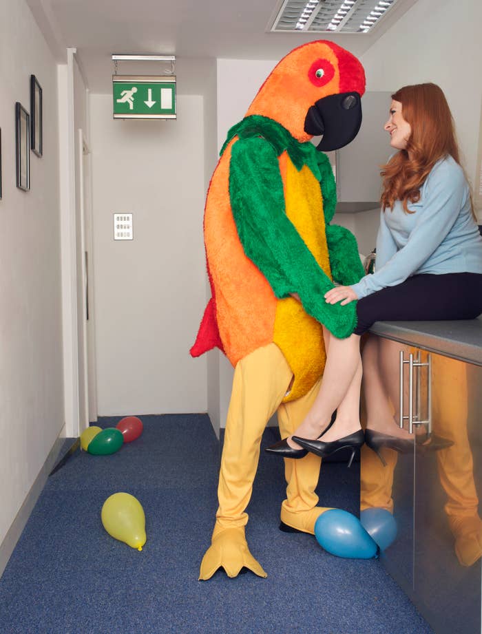 A person in a parrot costume interacts with a woman sitting on a countertop in an office-like setting with balloons on the floor