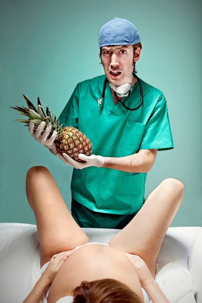 A doctor in scrubs and gloves, holding a pineapple, appears confused during a delivery with a pregnant woman lying down