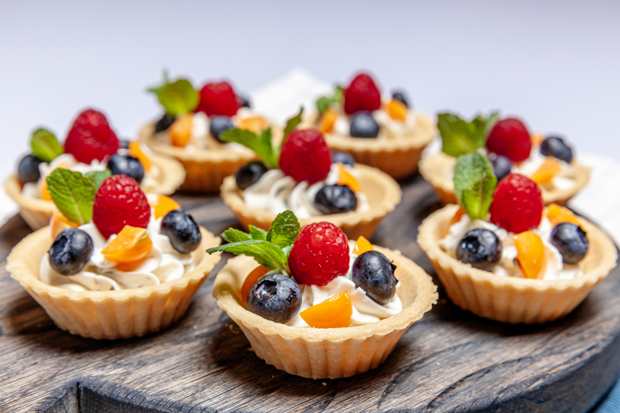Assorted fruit tarts with whipped cream, topped with raspberries, blueberries, orange segments, and mint leaves on a wooden surface