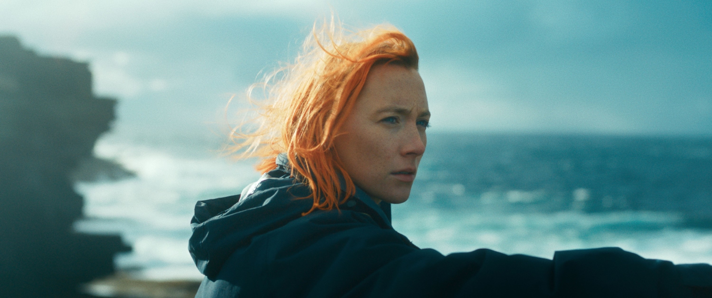 Saoirse Ronan stands on a rocky shoreline, gazing thoughtfully into the distance with windswept hair, wearing a jacket