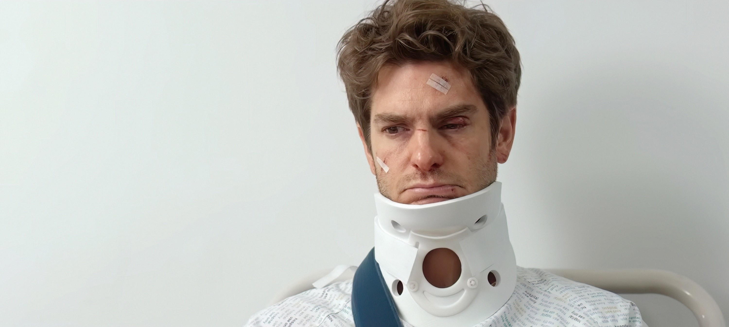 Andrew Garfield is seated with a neck brace and bandages on his face, appearing injured and solemn. He is wearing a hospital gown