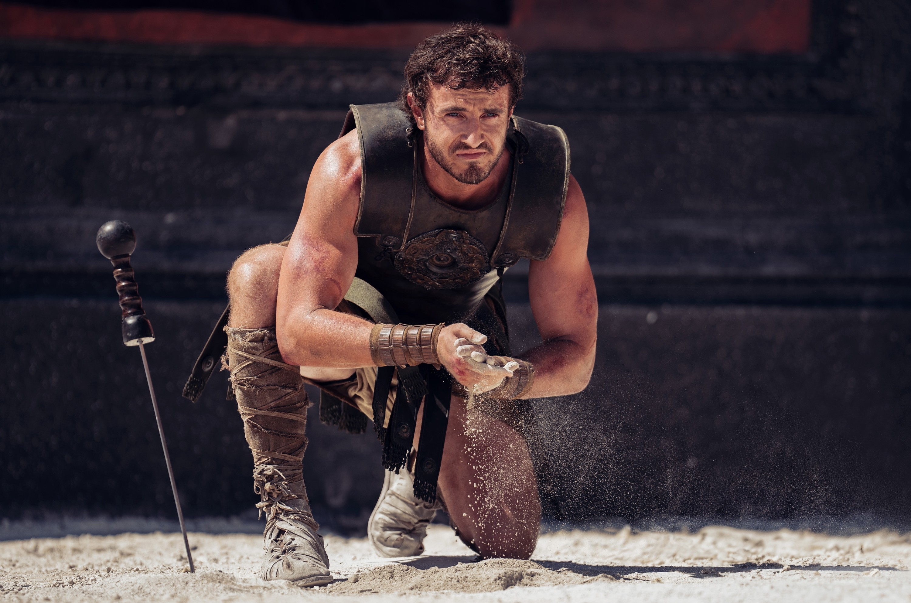 Actor kneels, wearing gladiator armor, gripping sand in one hand, with a sword planted in the ground beside him