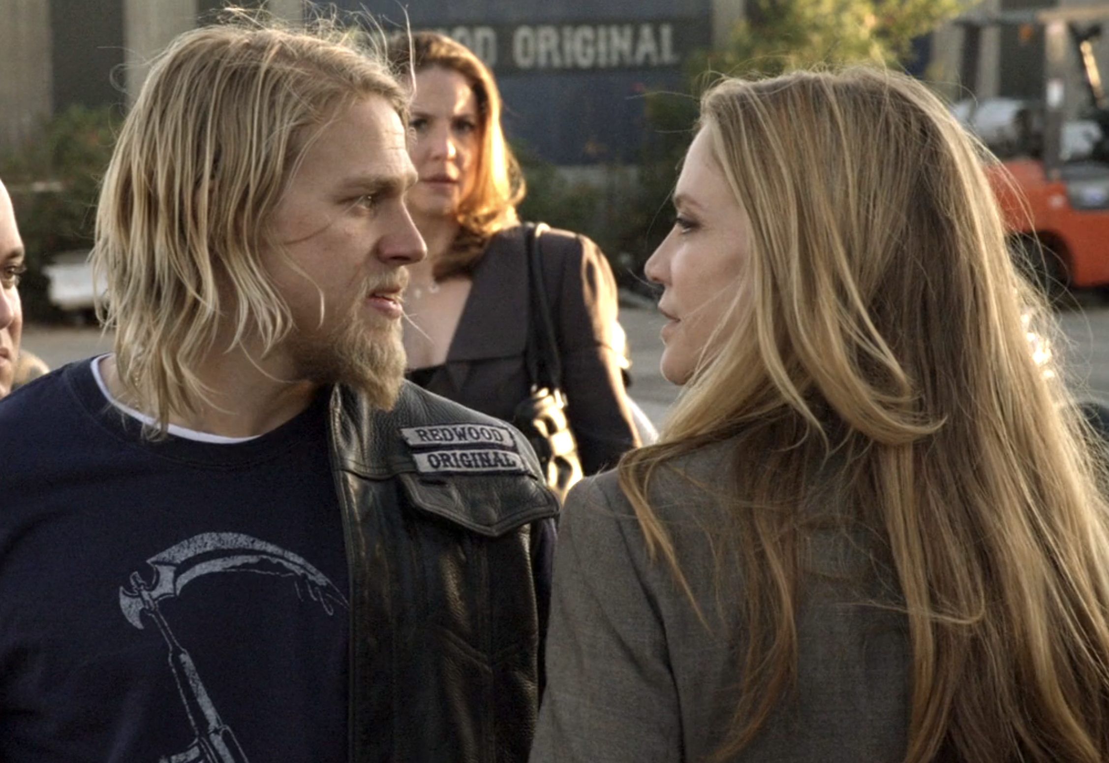 Charlie Hunnam and Winter Ave Zoli face each other in an outdoor scene. Hunnam wears a leather jacket with &quot;Redwood Original&quot; patch