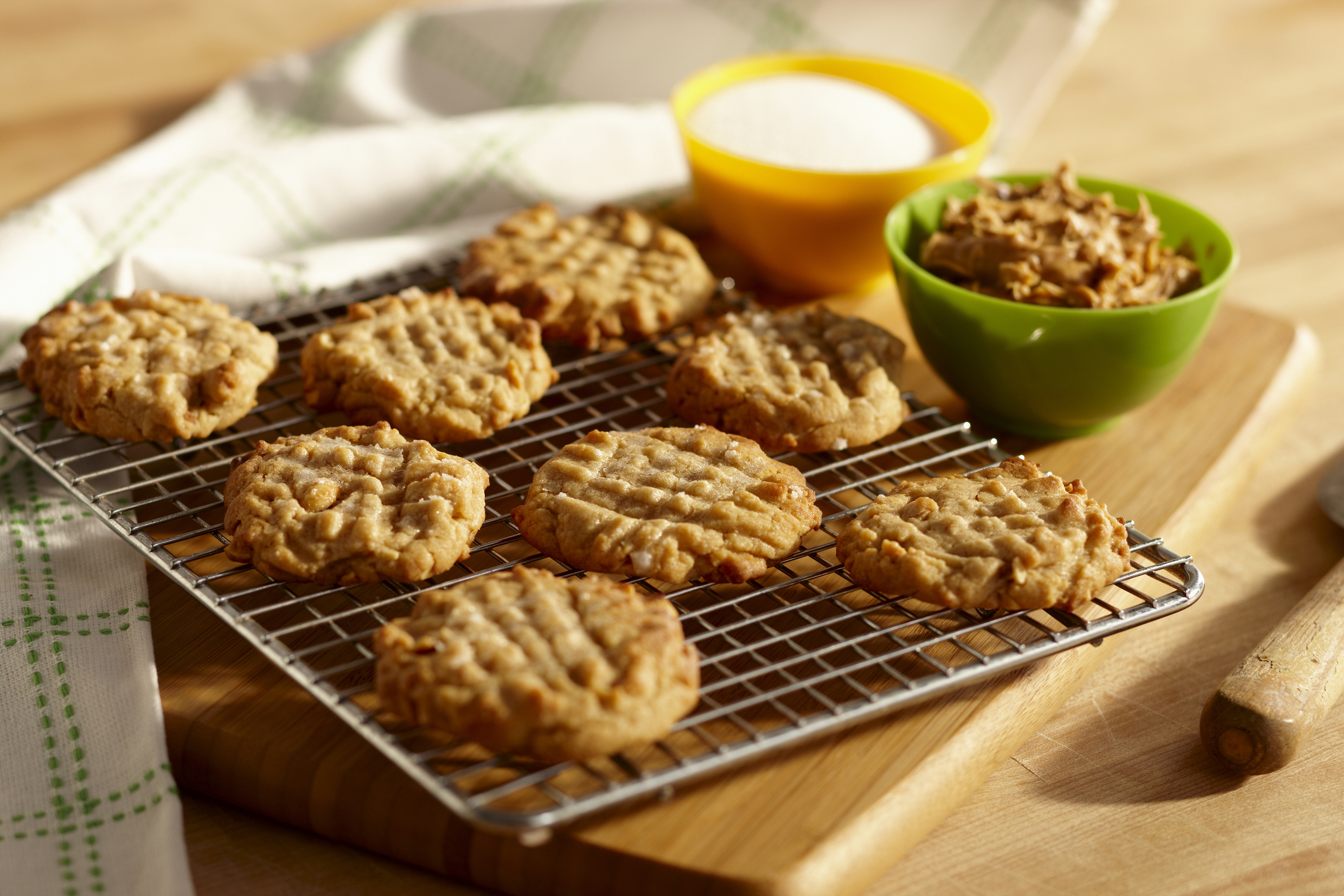Freshly baked peanut butter cookies on a cooling rack, with bowls of sugar and peanut butter in the background