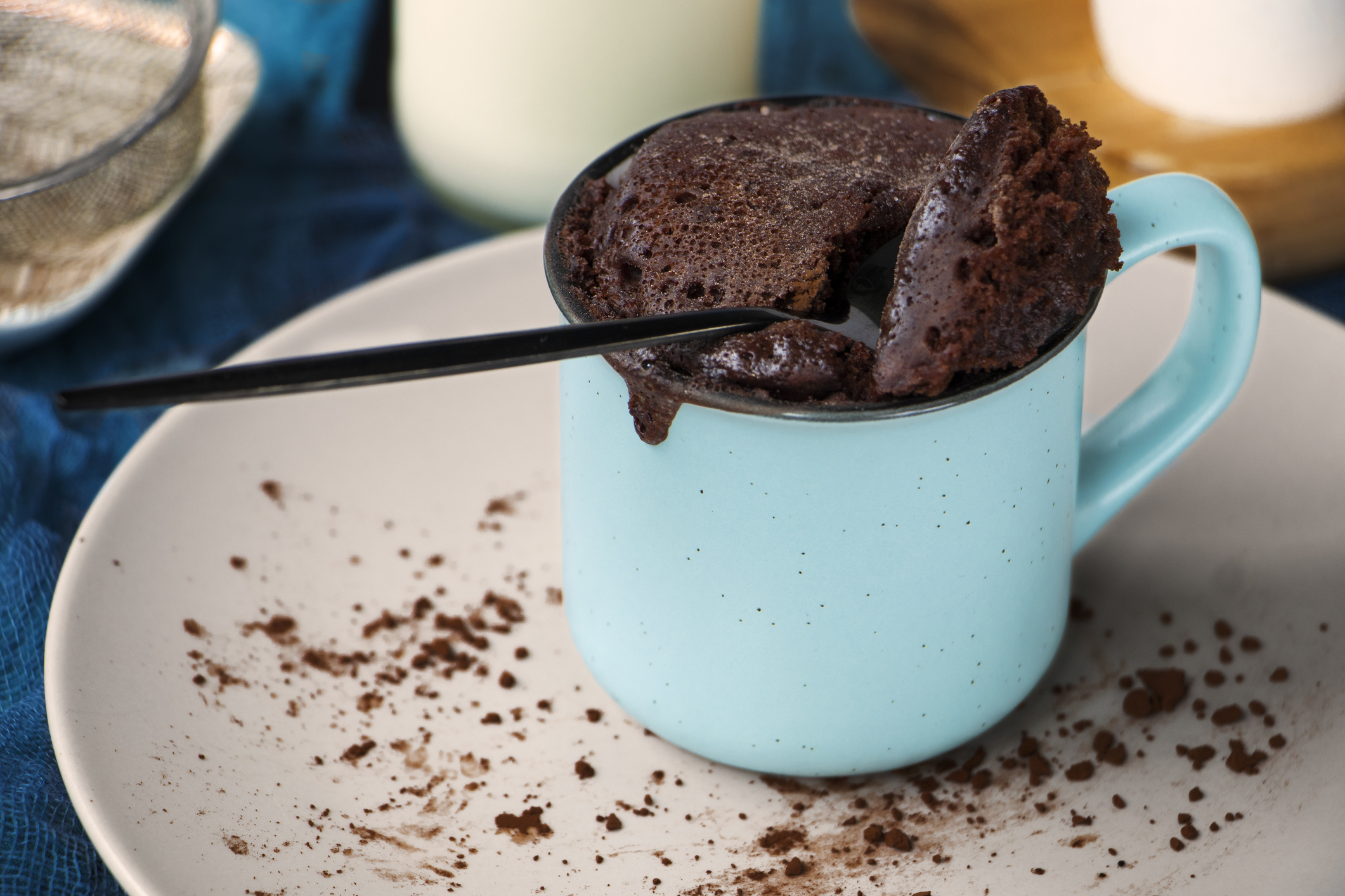 A chocolate mug cake in a blue mug with a spoon, placed on a plate with chocolate powder sprinkled around