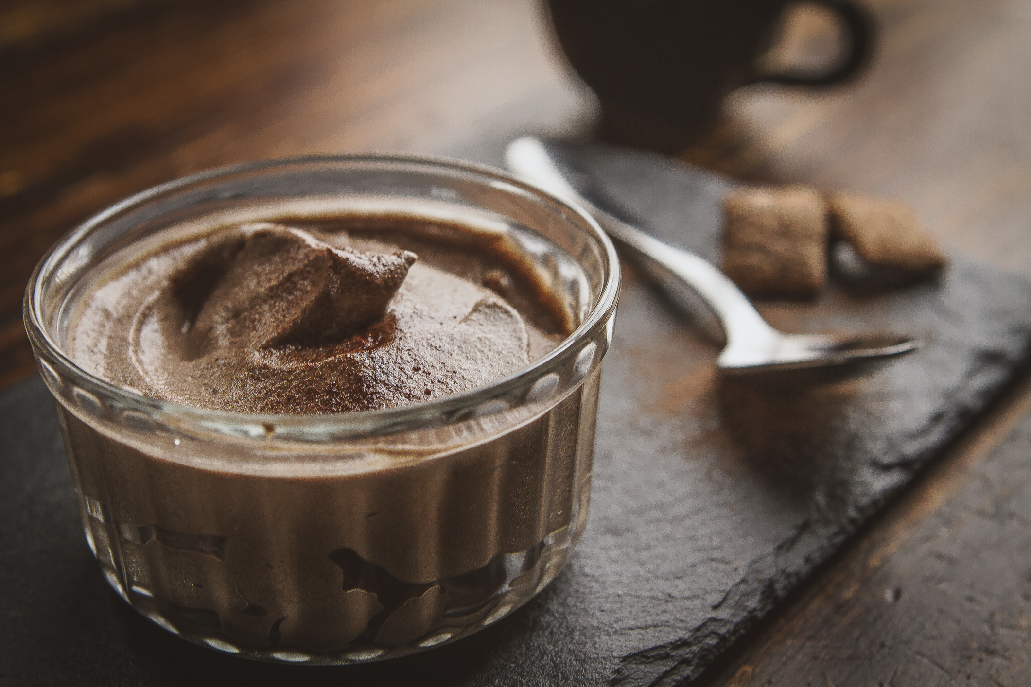 A glass dish filled with chocolate mousse sits on a dark surface next to a spoon and coffee cup