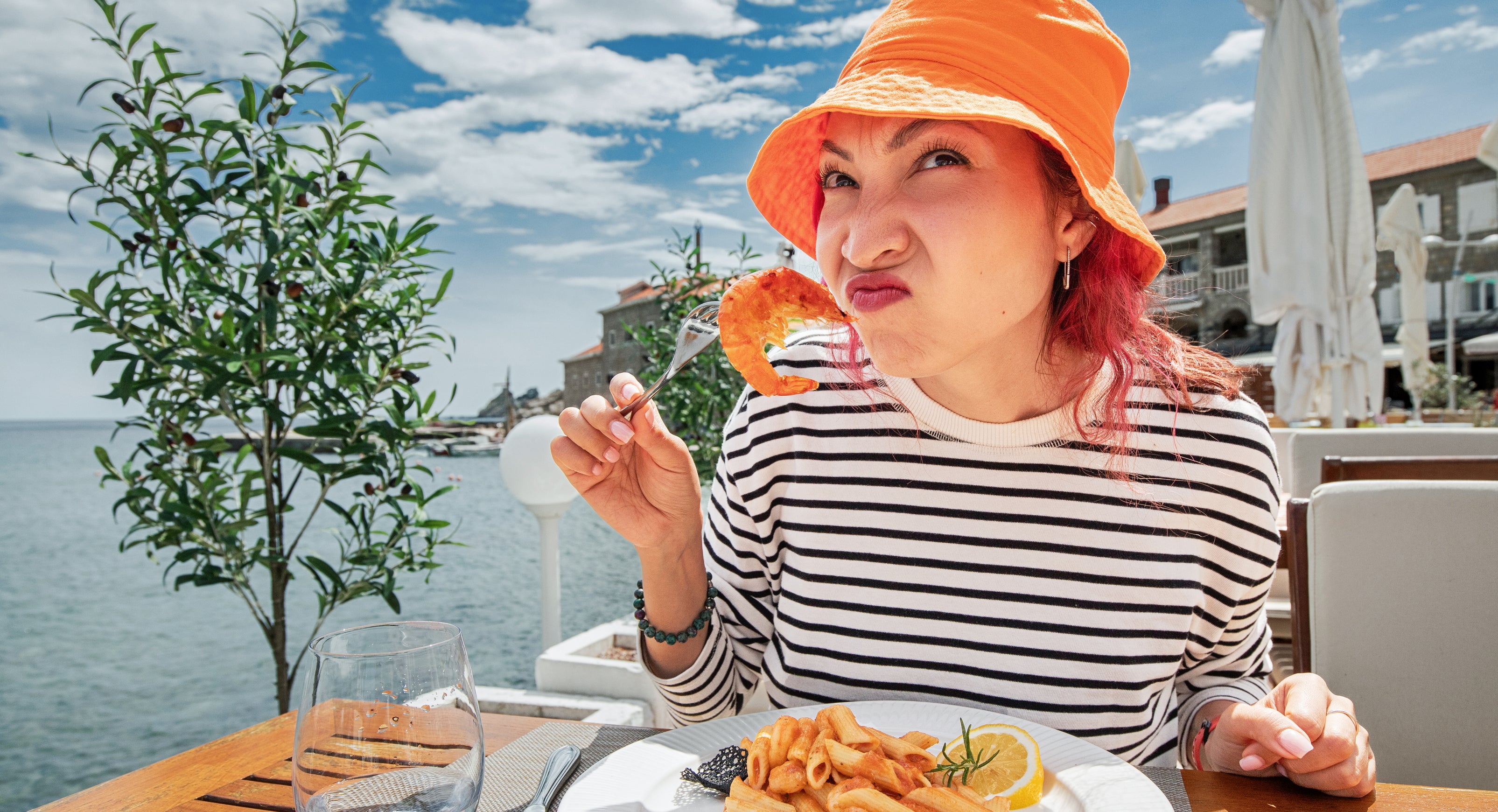 A person with red hair, wearing a bucket hat and striped shirt, eats seafood pasta at a waterfront restaurant, smiling playfully at the camera