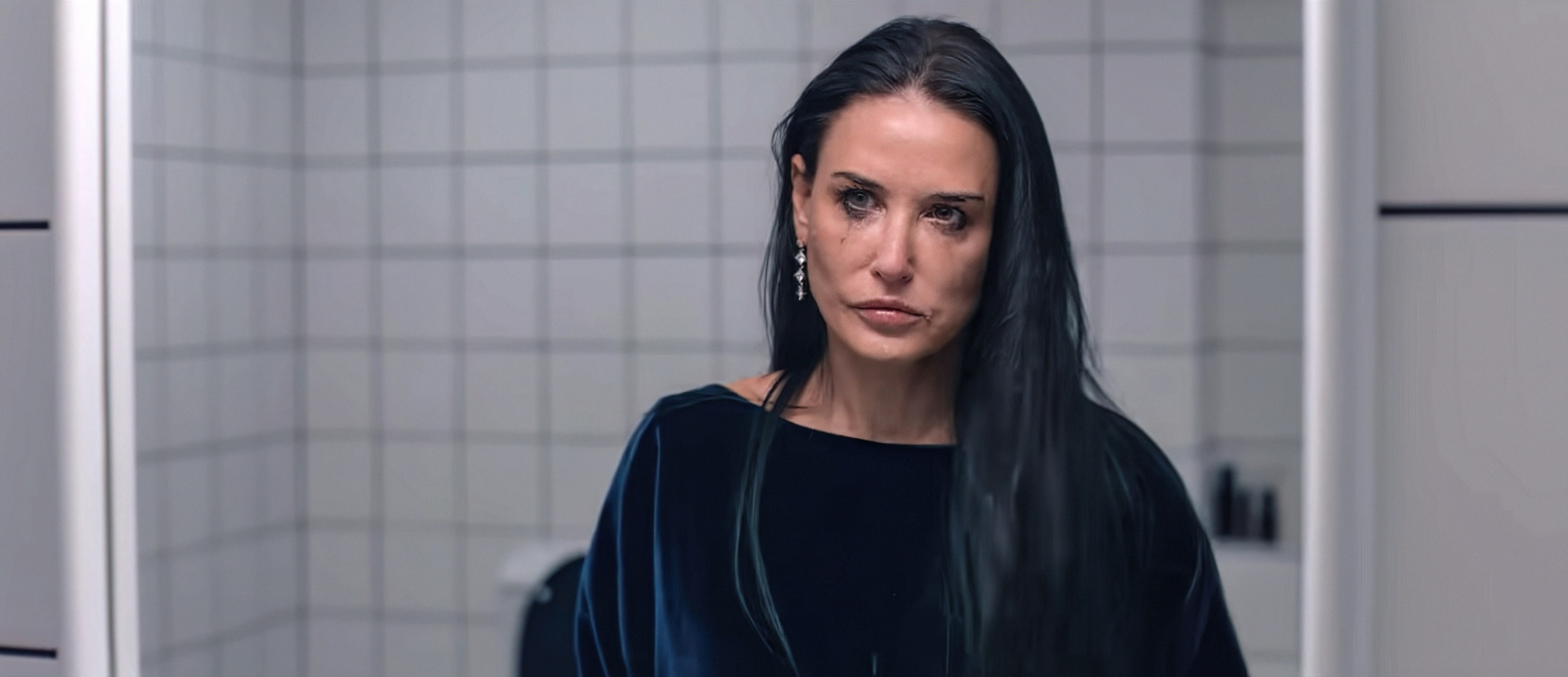 Demi Moore is standing in front of a tiled bathroom wall, looking directly at the camera with a somber expression. She is wearing a dark top