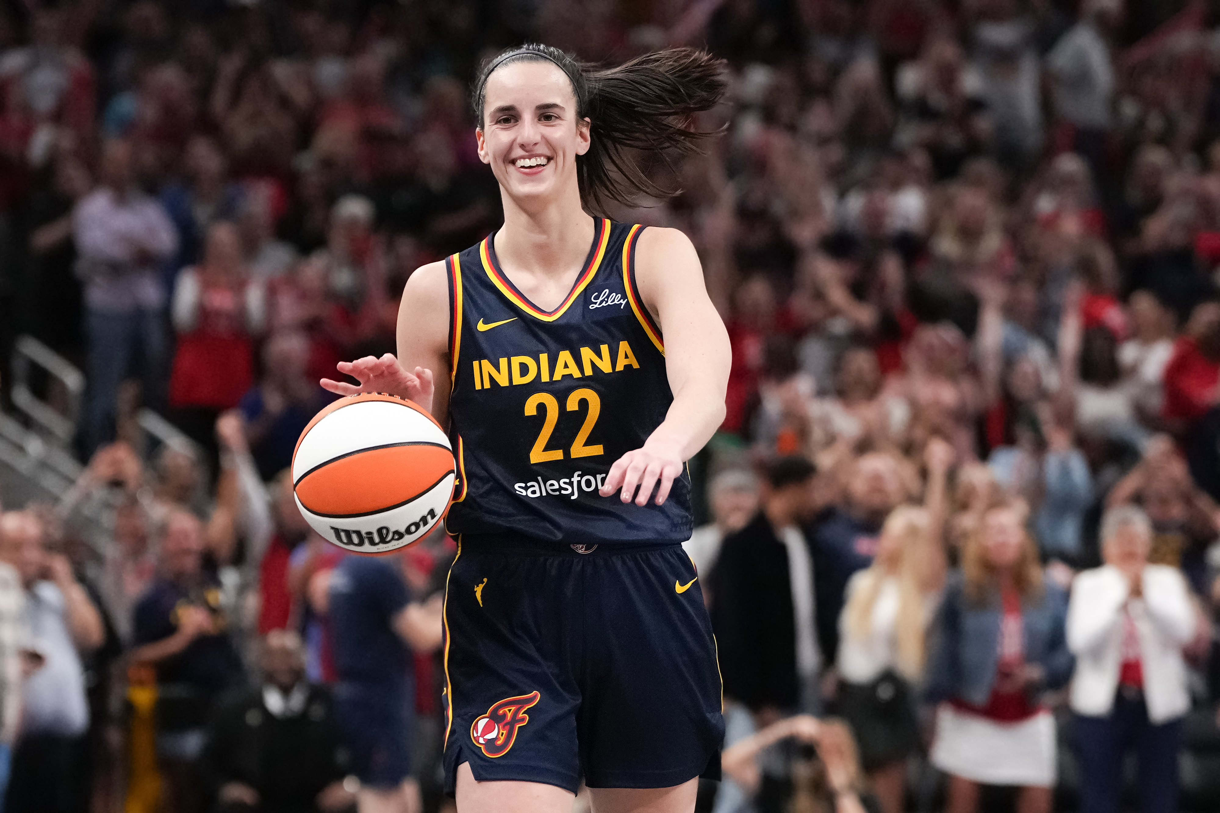 Aliyah Boston, in Indiana Fever uniform, holds a basketball on a crowded court, smiling widely as if celebrating a moment in a game