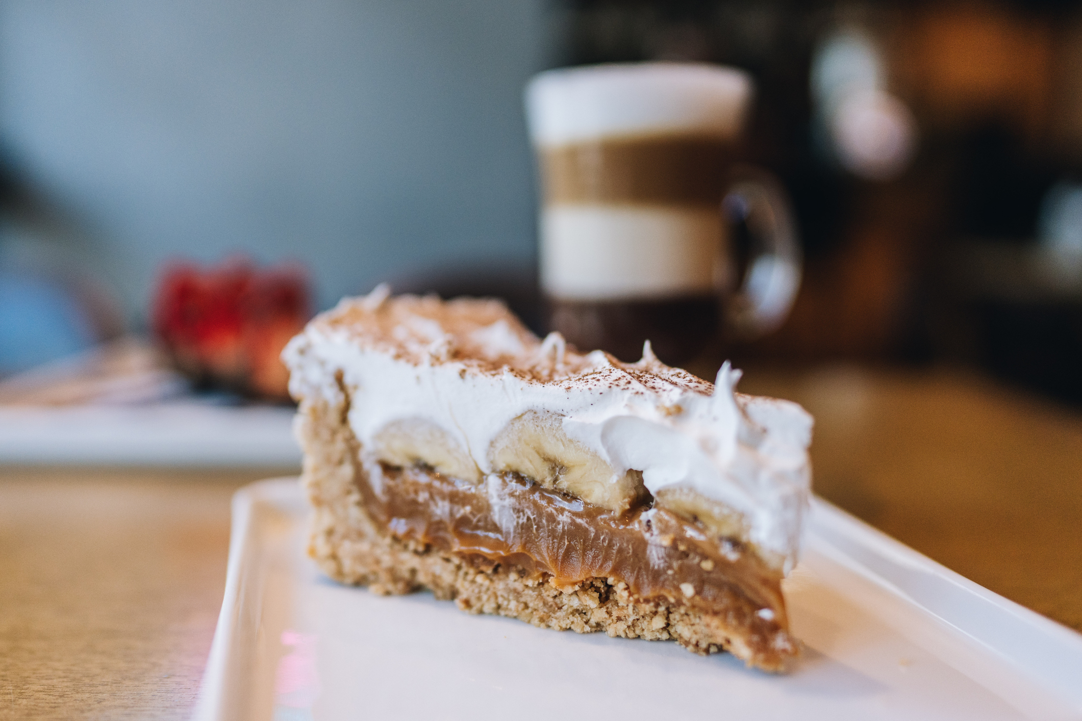 A slice of banana cream pie with layers of cream, bananas, and caramel on a plate. A layered coffee drink is in the background