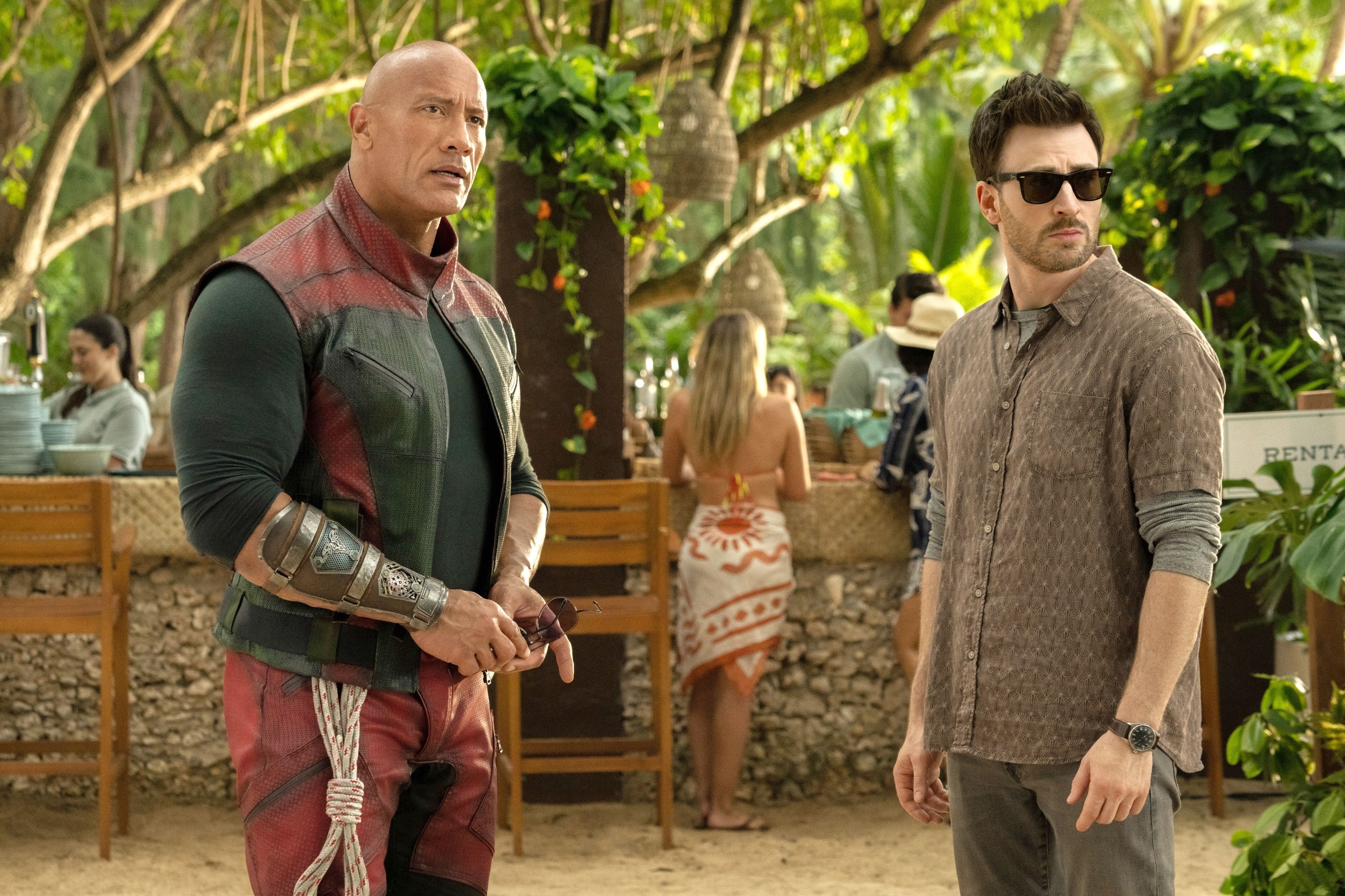 Dwayne Johnson in a futuristic outfit and Chris Evans in a casual shirt and sunglasses on a film set. People are in the background near a bar