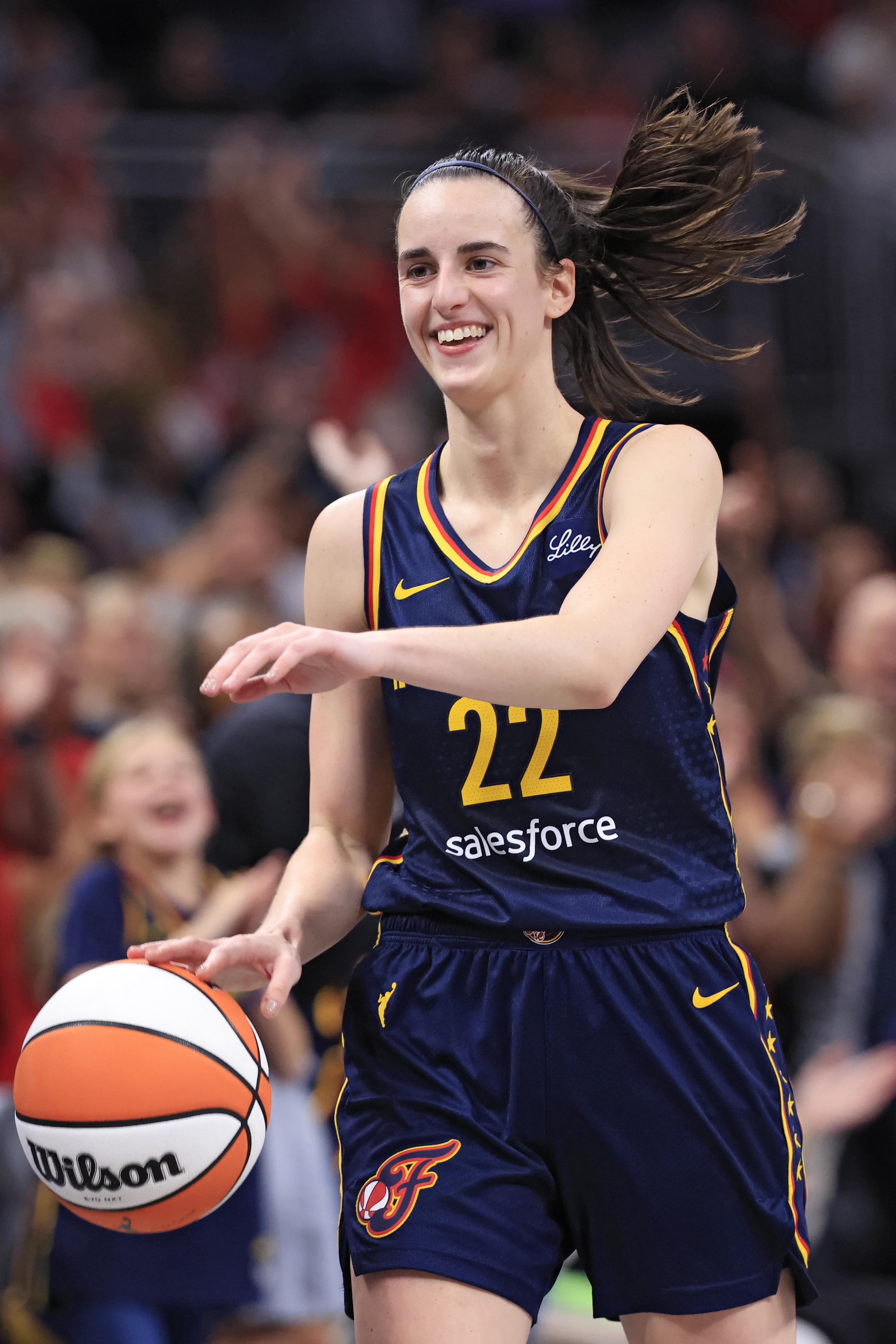 Caitlin Clark playing basketball, wearing a jersey with number 22 and the name "Lilly." She is smiling and holding a Wilson basketball