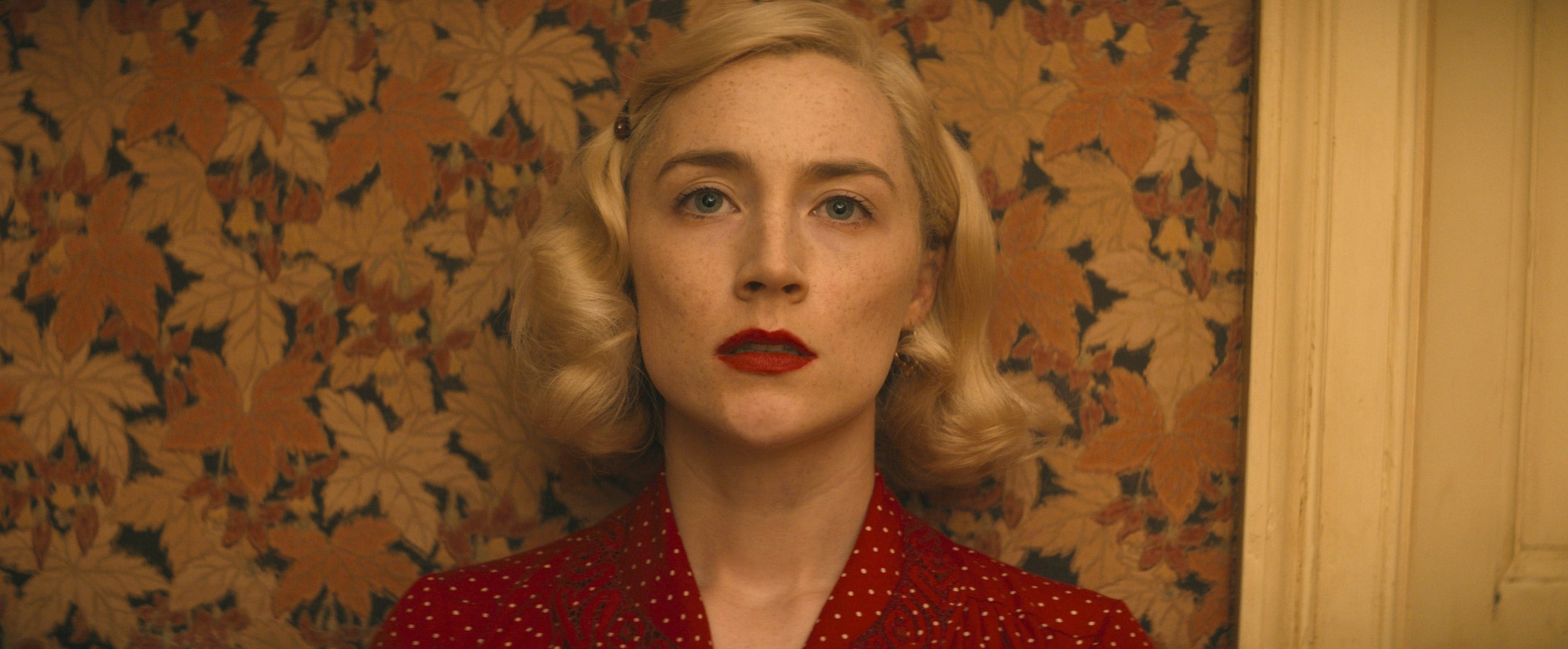 Saoirse Ronan in 1940s-style attire with wavy blonde hair and red lipstick, standing against a vintage leaf-patterned wallpaper