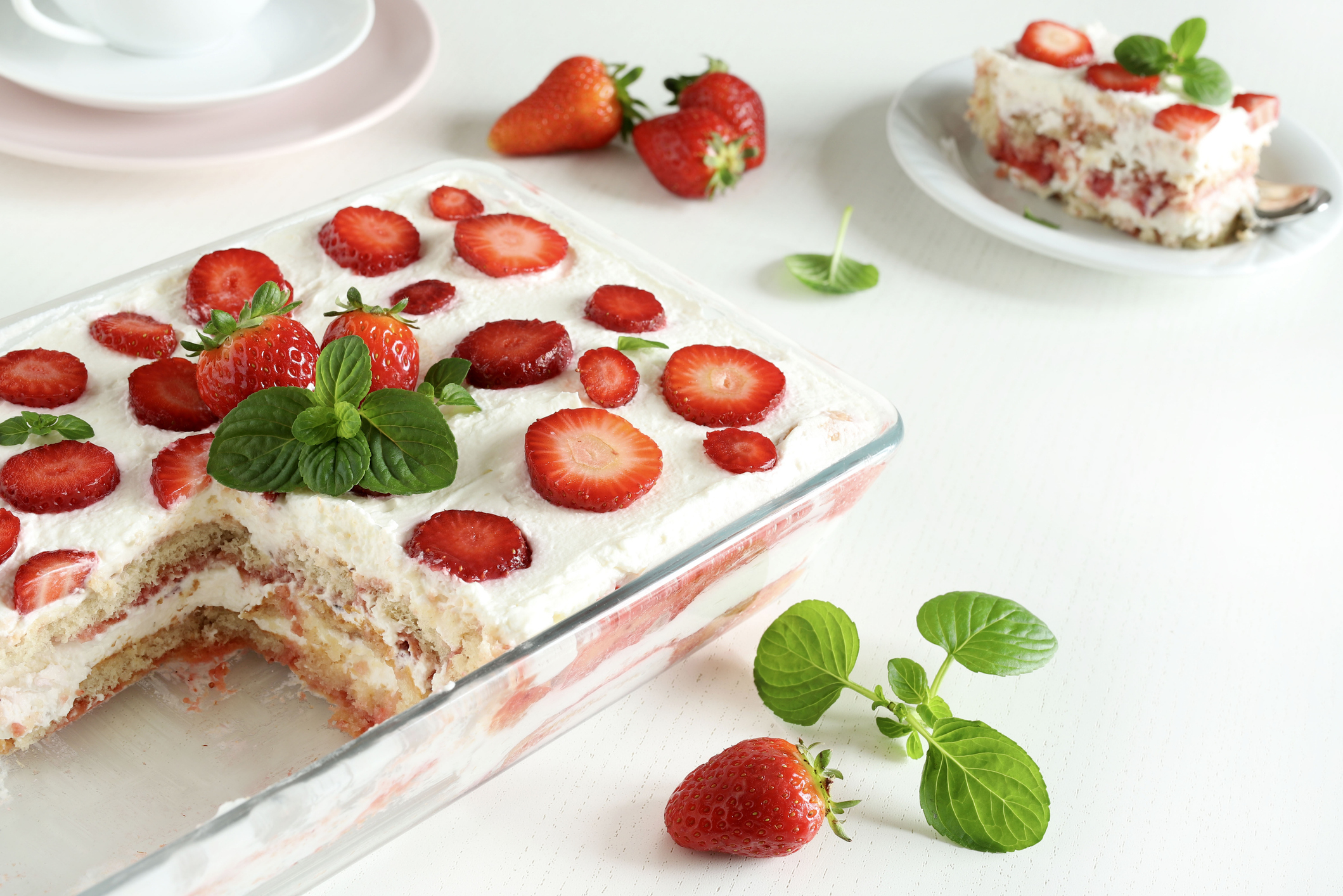 Strawberry tiramisu in a glass dish with a piece served on a plate nearby. Fresh strawberries and mint leaves are placed around the dishes on a white table
