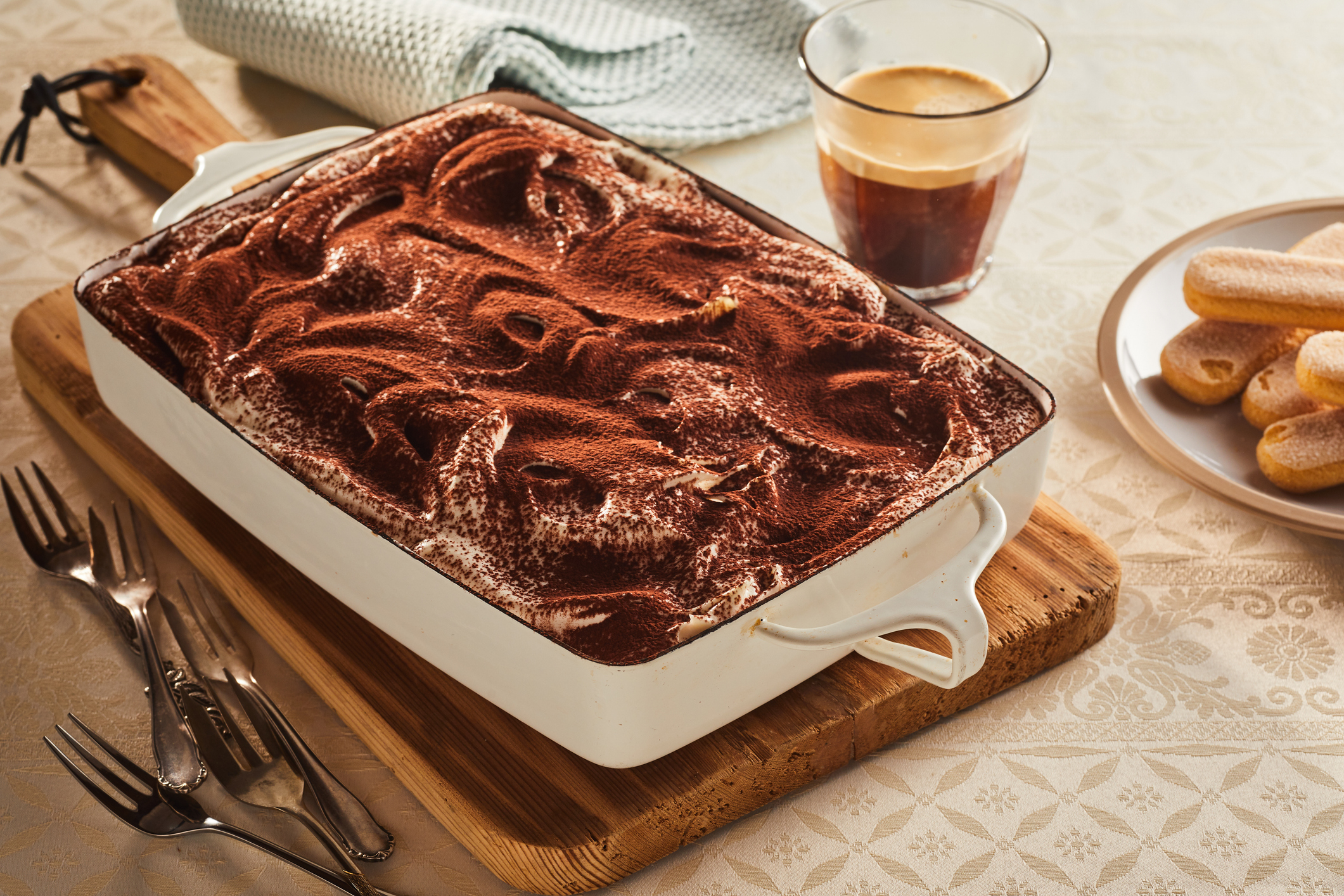 Tiramisu in a white dish on a wooden board with a glass of espresso and ladyfingers on the side. Four forks lie nearby on a tablecloth