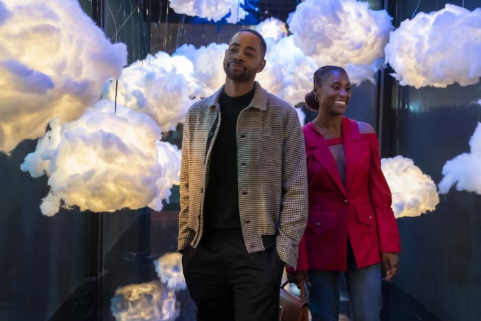 Issa Rae and Jay Ellis walk through an art installation with cloud-like sculptures. Jay is in a casual jacket and dark pants, and Issa in a stylish blazer