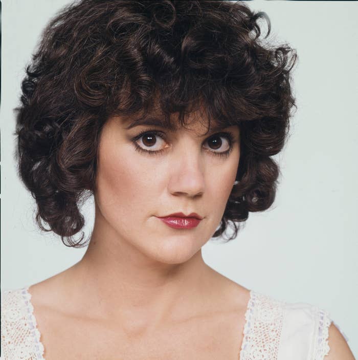 Woman with curly, short hair posing with a serious expression, wearing a lace-trimmed white top. No additional identifiable individuals present