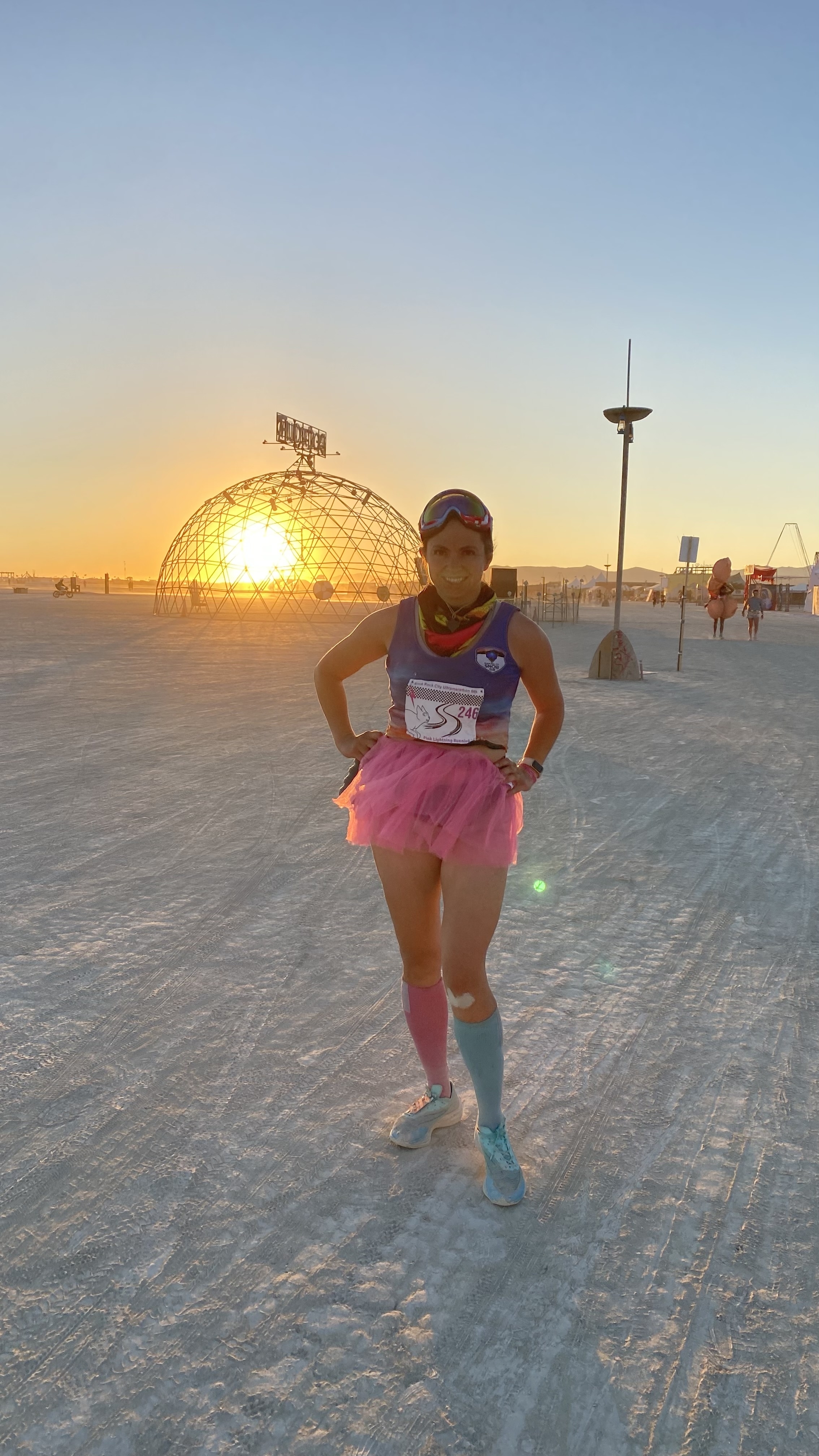 Elizabeth standing outdoors on a vast, open area at sunset, wearing a pink tutu, blue and pink socks, and running gear. The sun is setting behind a dome structure